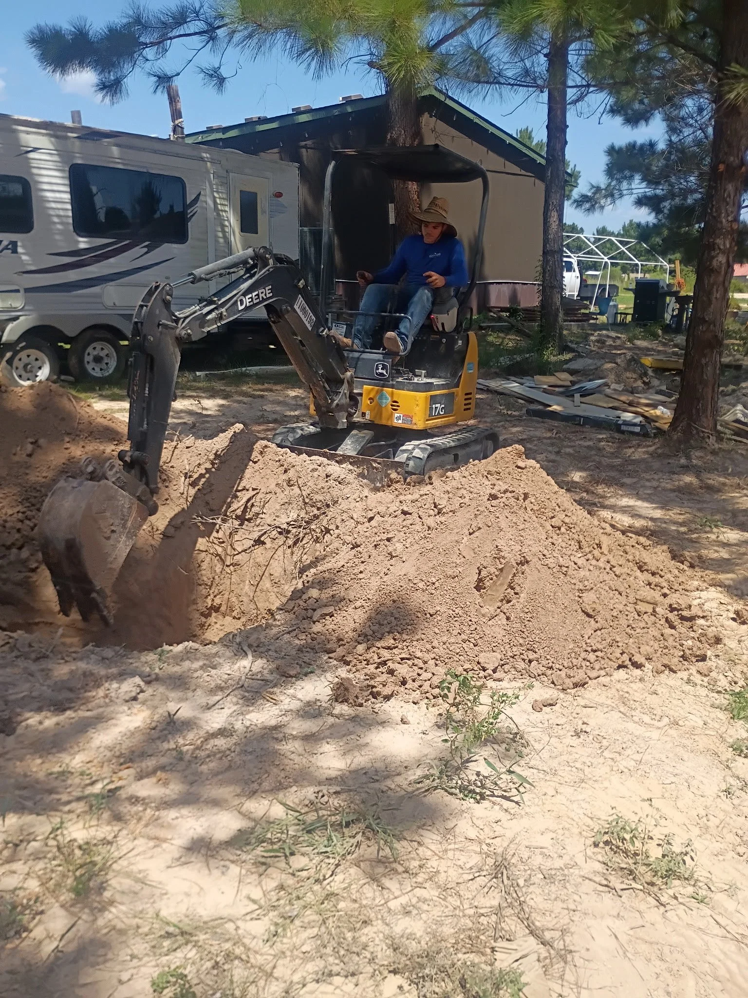 A person operating a small John Deere excavator on a construction site with dirt, trees, and some construction materials and vehicles in the background.