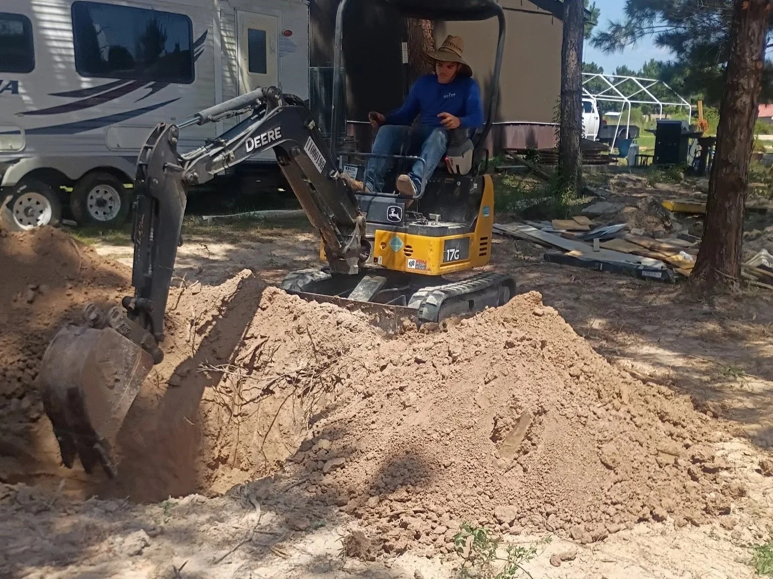 A woman operating a small John Deere excavator digging a hole in sandy ground outdoors.