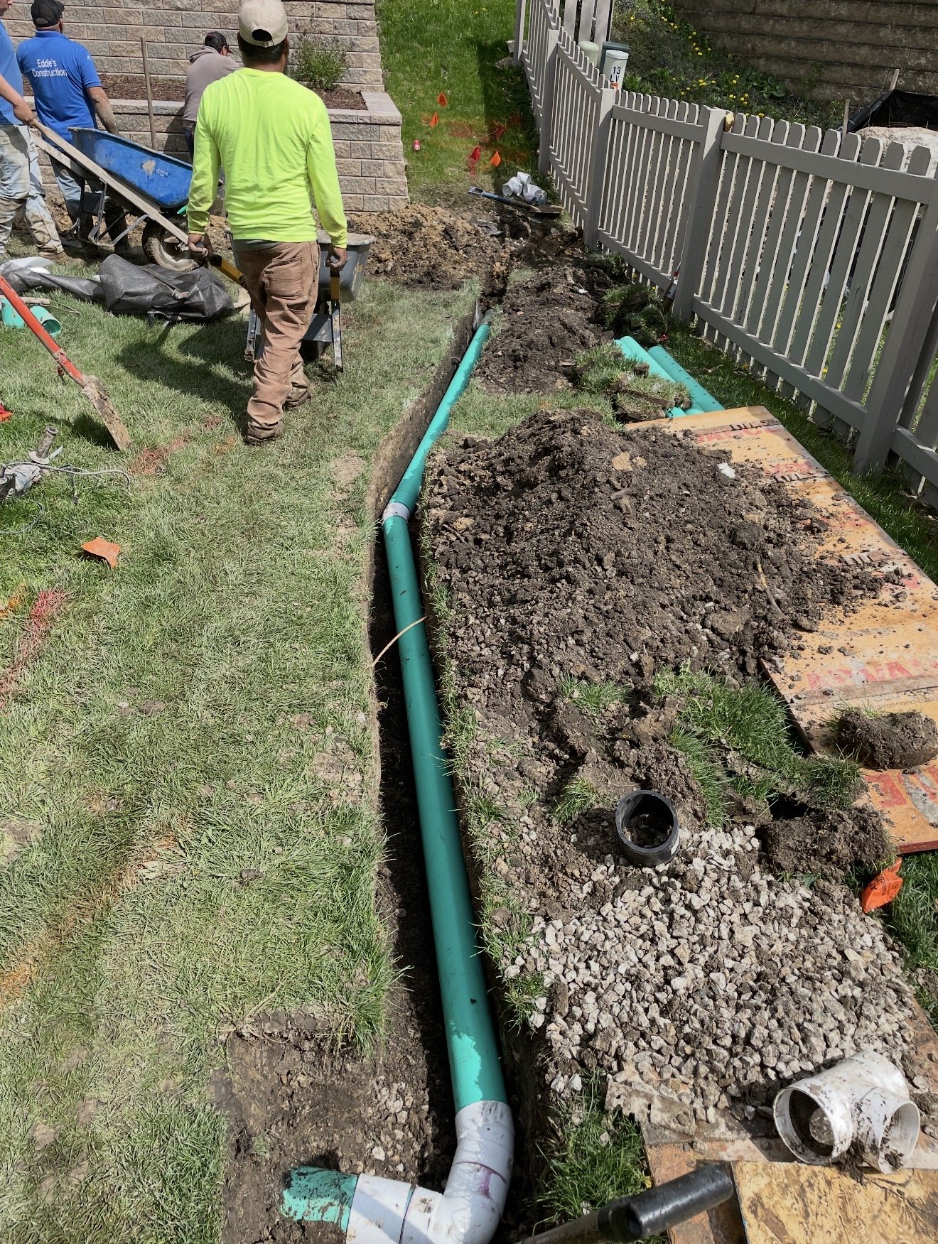 Workers install underground piping along a trench in a backyard, with tools and construction materials nearby.
