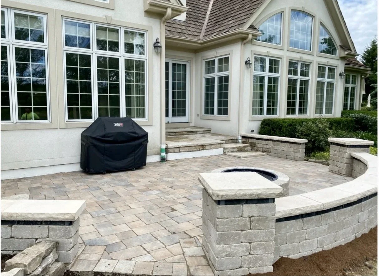 Backyard patio with stone pavers, built-in fireplace, a barbecue grill covered with a black cover, and large house windows with a door and steps leading to the house interior.