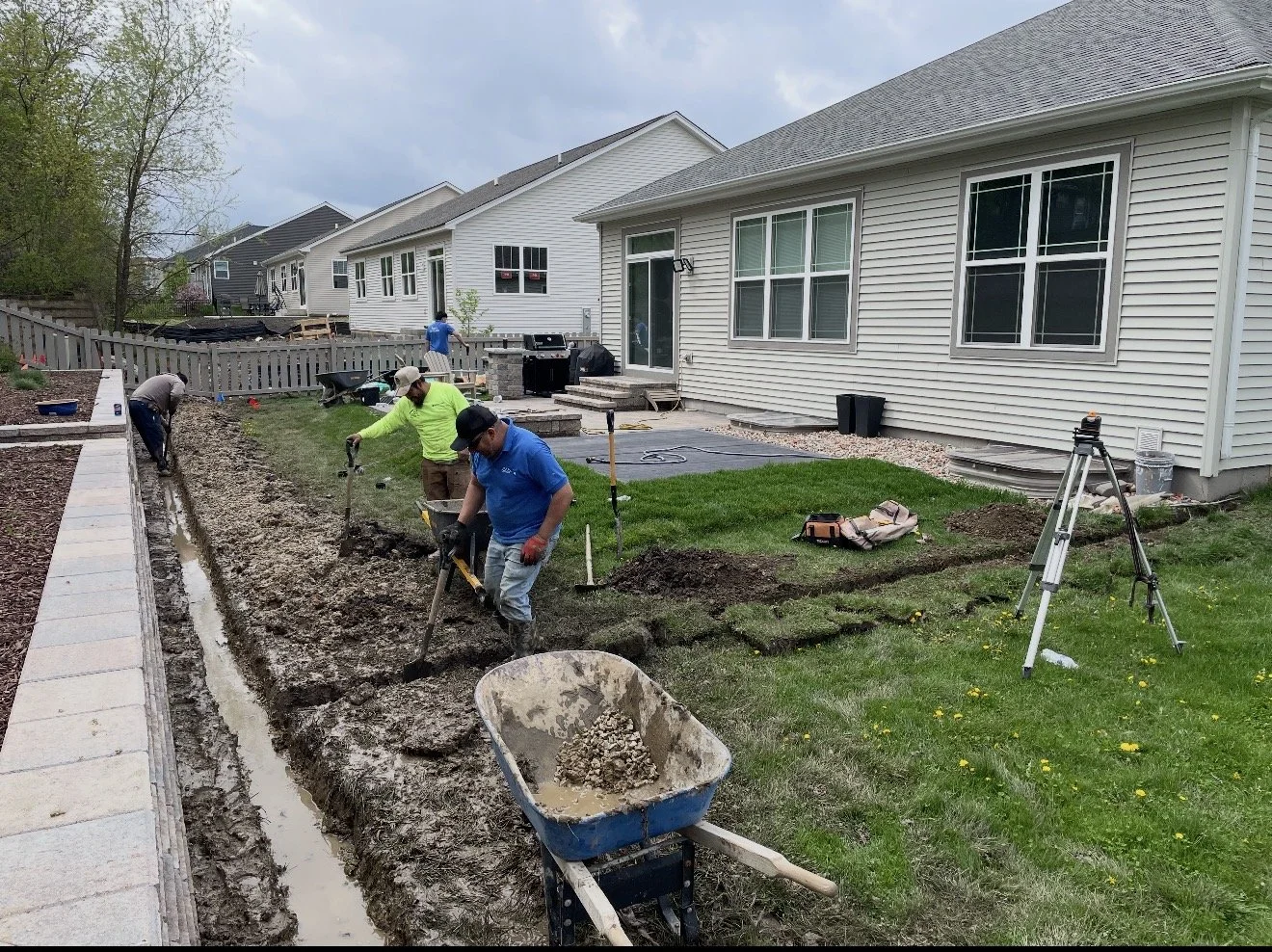 People working on a landscaping project in a backyard, using shovels and tools to prepare the ground for paving or planting, with a wheelbarrow in the foreground and a house with a fenced yard in the background.