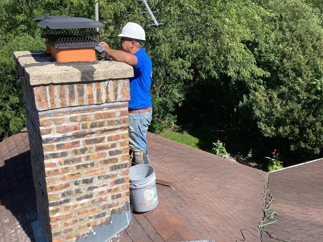 A worker repairing a brick chimney on a residential roof, wearing a white safety helmet, blue shirt, jeans, and boots, with a bucket nearby and green trees in the background.