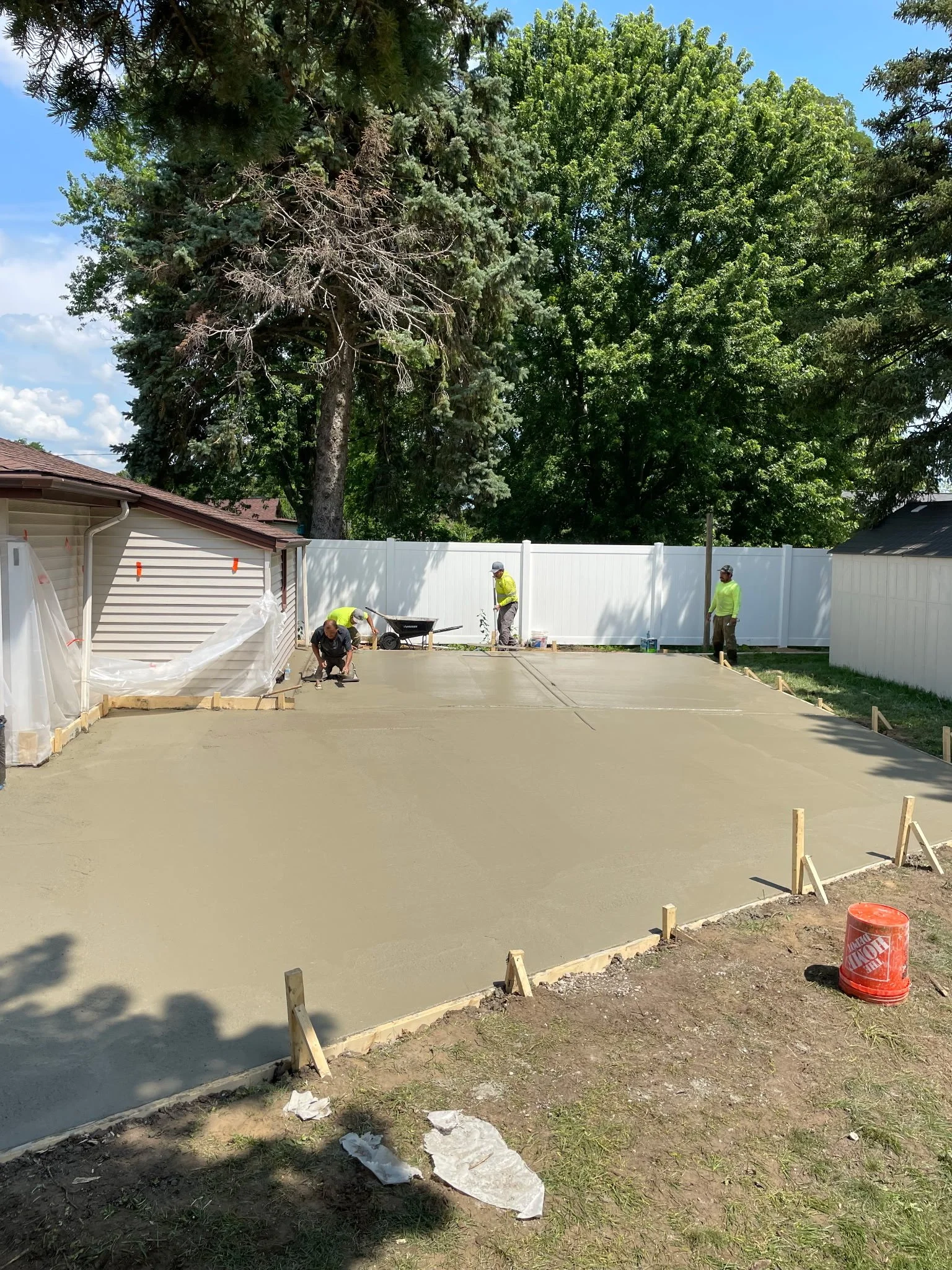 Workers applying fresh concrete for a driveway in a residential backyard, with a white fence, large trees, and a house in the background.