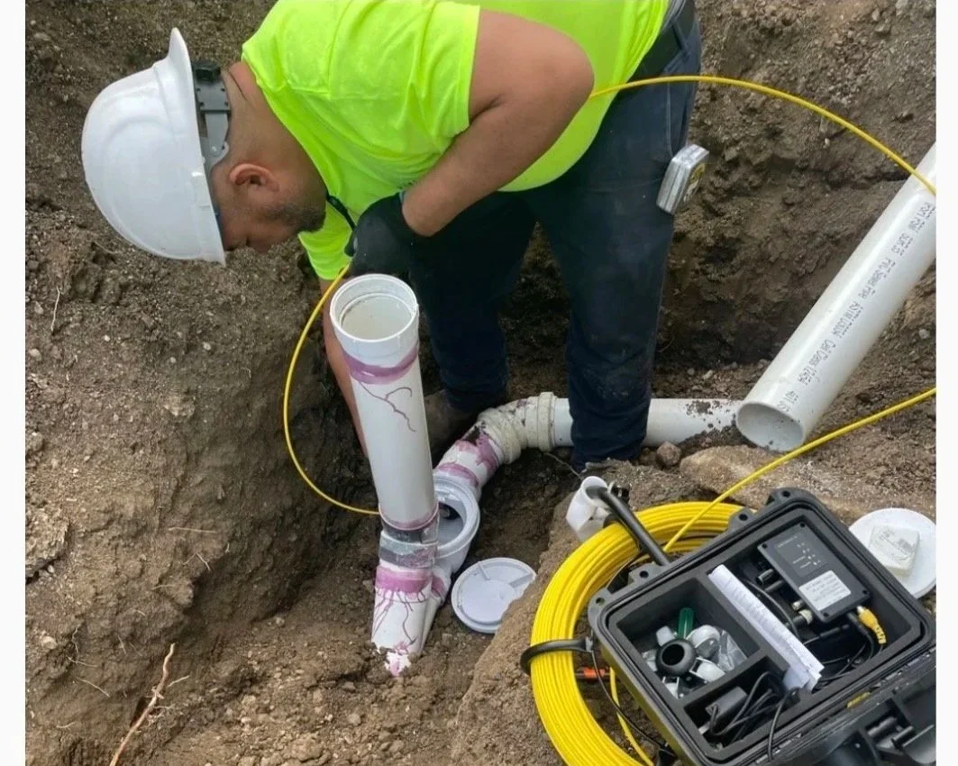 A worker in a yellow safety vest and white hard hat crouches in a trench, working on underground piping and electrical cables. Various tools and pipes are around him.