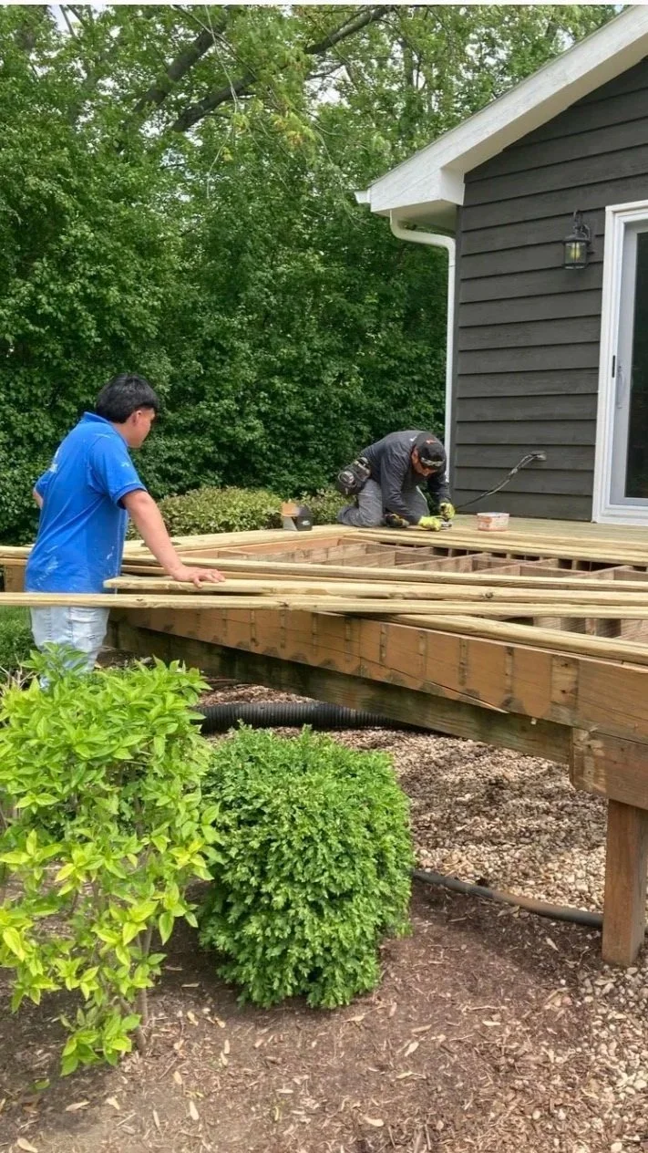 Two men are working on building a wooden deck outside a house. One man, wearing a gray shirt and cap, is kneeling and using tools, while the other man, in a blue shirt, is standing and observing. The deck is above ground level, supported by wooden posts, and there are trees and bushes nearby.