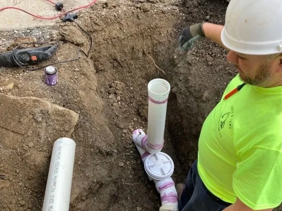 A construction worker wearing a white hard hat and bright yellow shirt is working on underground plumbing, installing PVC pipes in a dug trench.