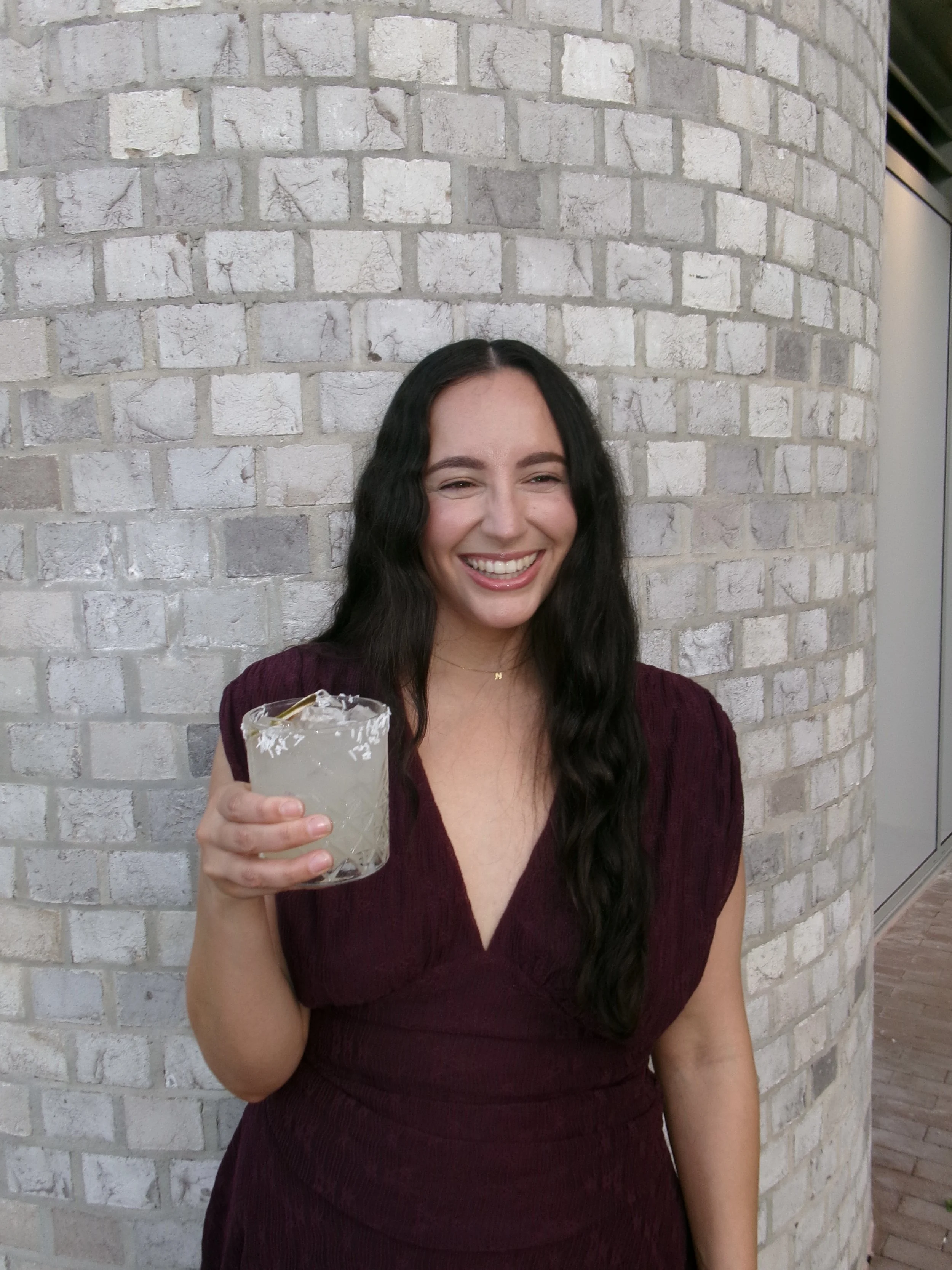 A woman with dark hair smiling and holding a margarita glass with a salted rim in front of a white brick wall.