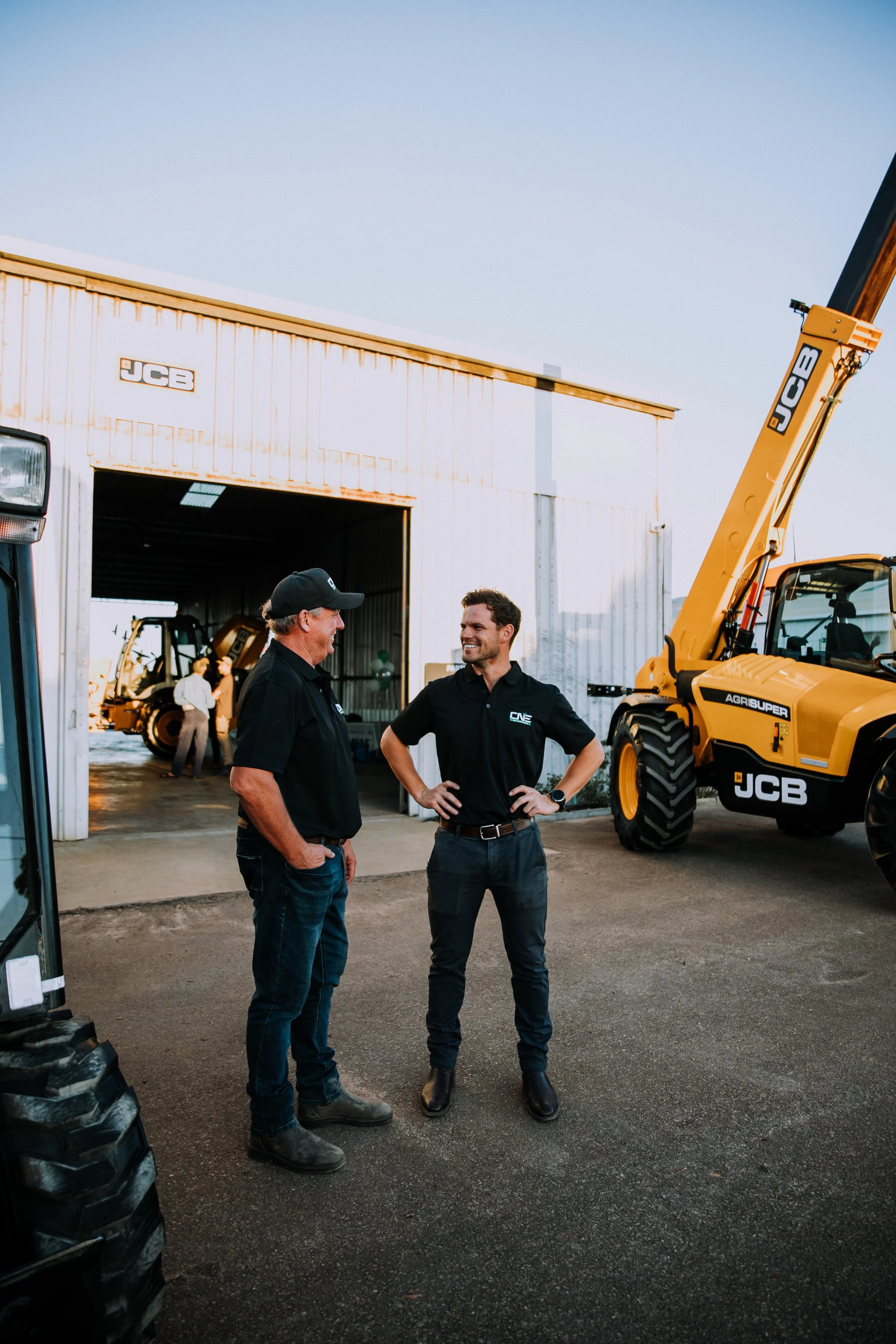 Two men talking outside a JCB dealership shed with telehandlers on display