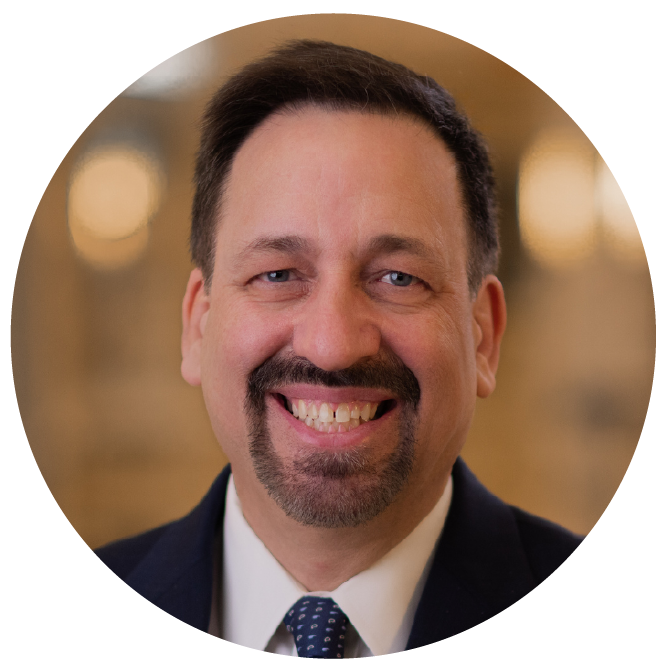 Smiling man with a beard and mustache, wearing a suit and tie, in an indoor setting with warm lighting.