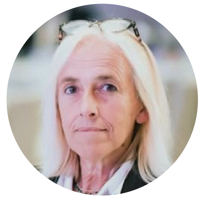 Close-up of an older woman with long white hair, wearing glasses on her head, and a black blazer, looking slightly to the side.