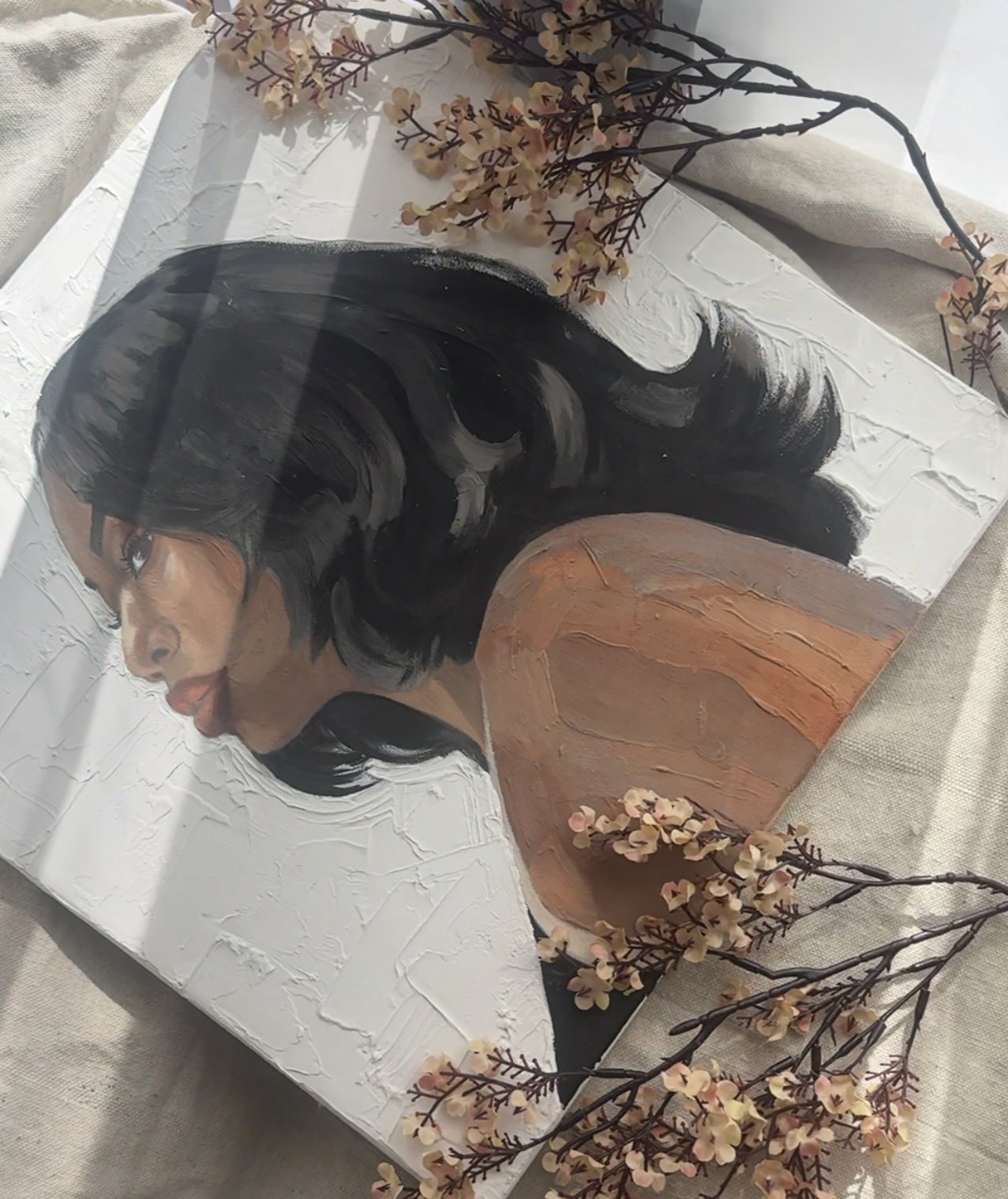 A painting of a woman with black hair, gazing to the side with her tongue slightly out, featuring textured white background and decorative branches with small pink flowers surrounding it.