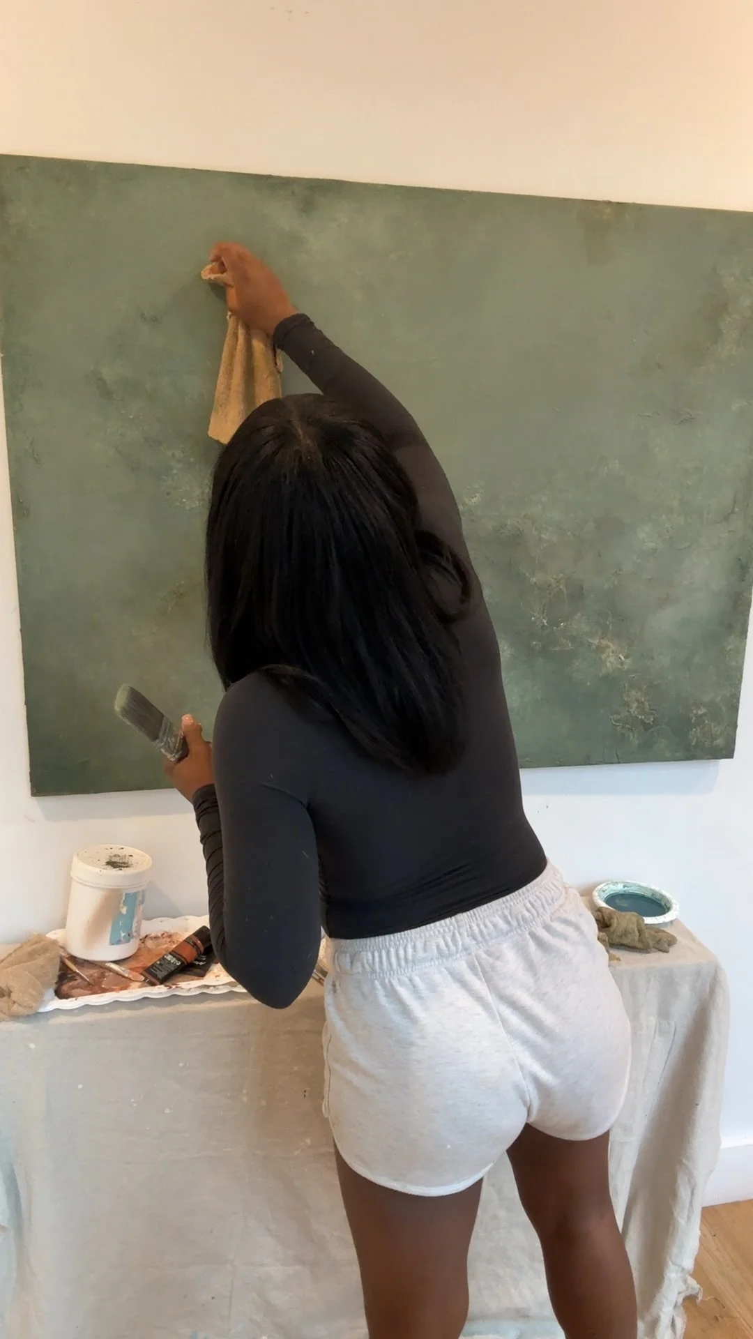 Person painting a textured wall, wearing a black long-sleeve shirt and white shorts, holding a paintbrush and a container of paint, with painting supplies on a nearby table.