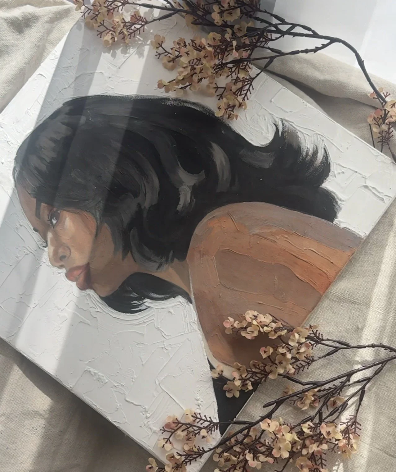 Painting of a woman with black hair, wearing a brown top, depicted from the side. The painting has a textured white background with floral embellishments.