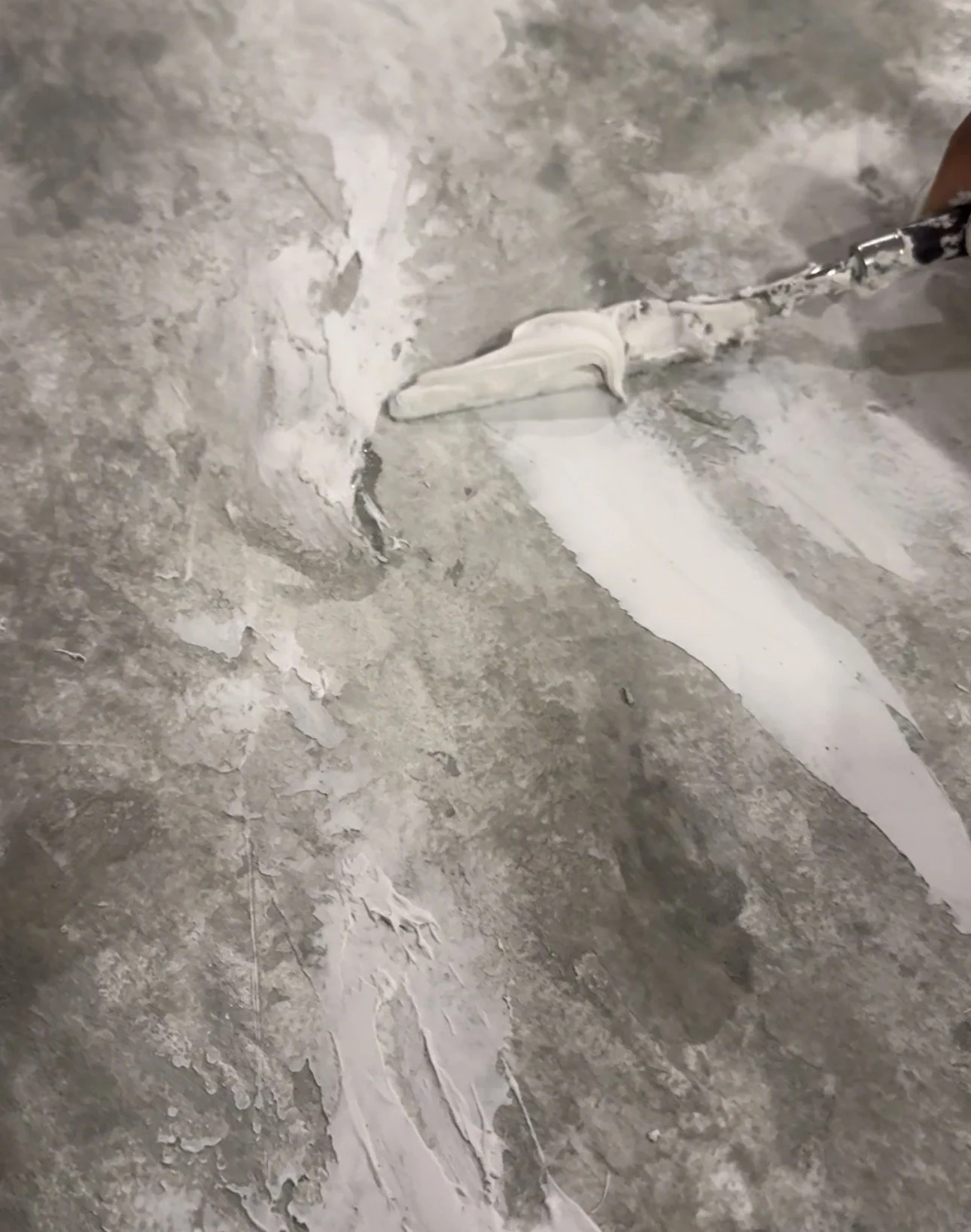 Person applying white floor adhesive or compound with a trowel on a concrete or tiled floor.