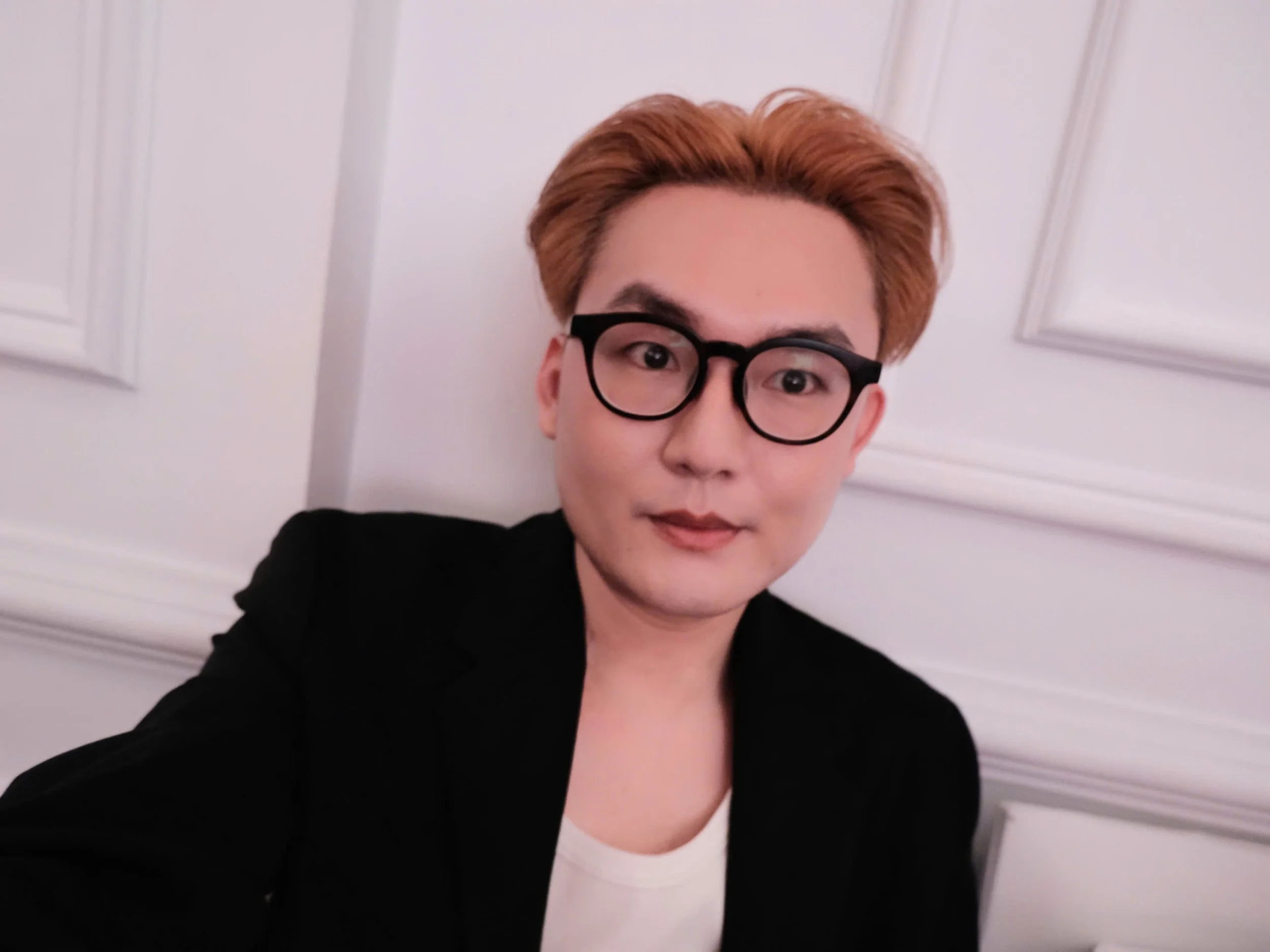 Close-up of a person with light skin, reddish-brown hair, black glasses, wearing a black jacket and a white shirt, in front of a white wall with molding details.