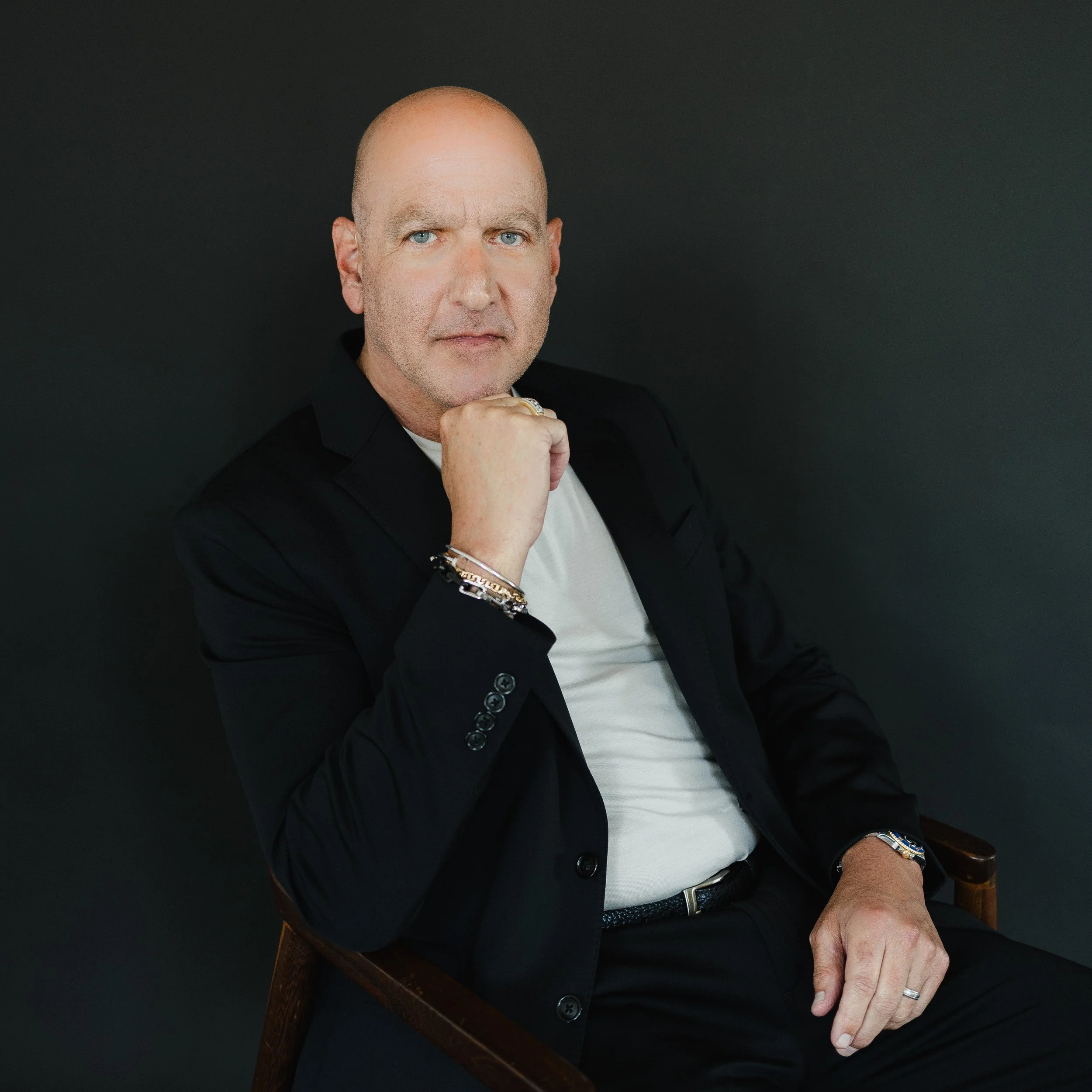 A close-up image of a bald man with blue eyes sitting against a plain dark background. He is wearing a black jacket, a white shirt, and accessories including a watch and bracelets. His left hand is resting on his chin, and he is looking directly at the camera.