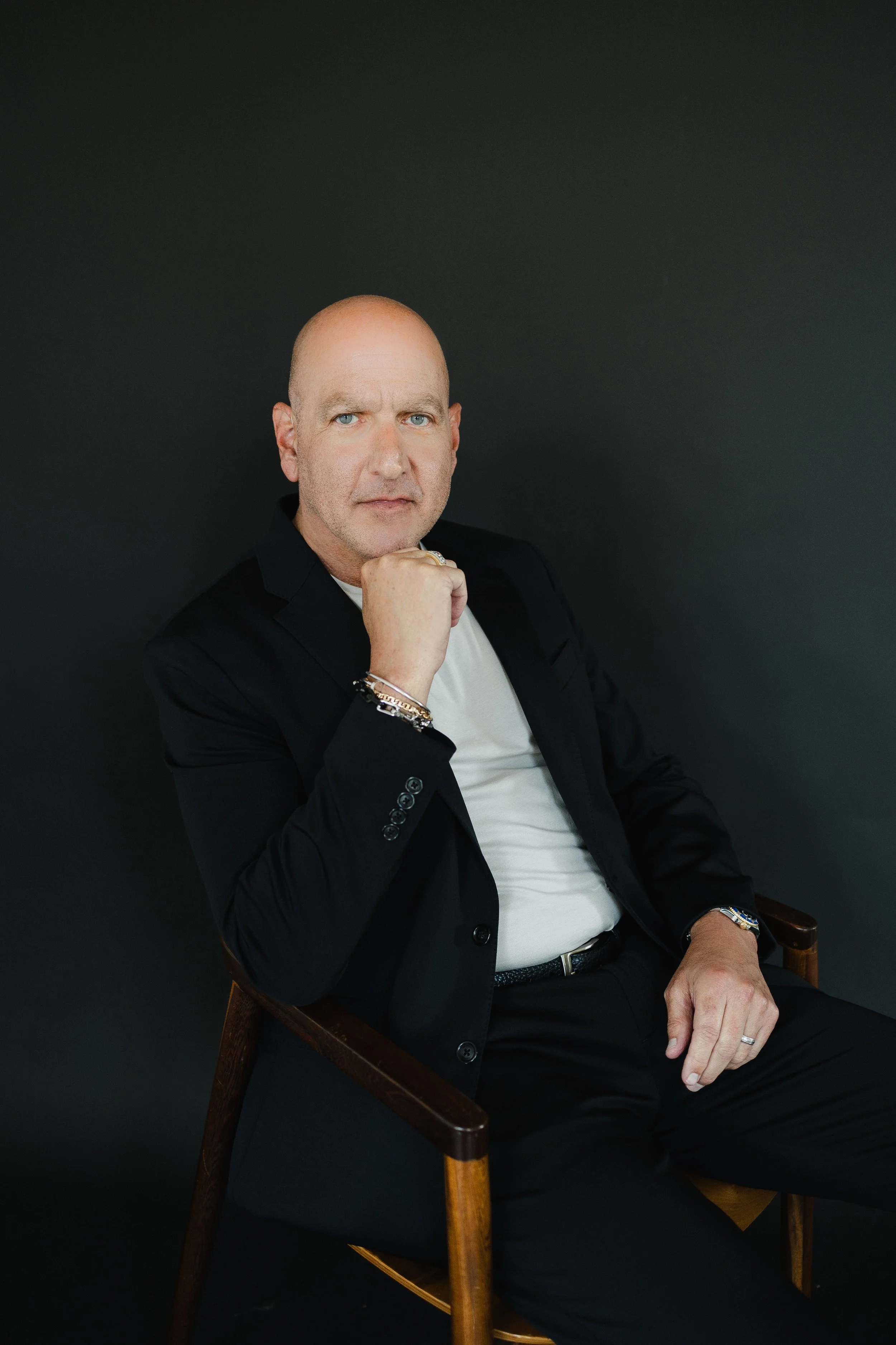 A middle-aged man with blue eyes and a bald head, dressed in a black blazer and white shirt, sitting on a wooden chair against a dark background, resting his chin on his hand while looking at the camera.