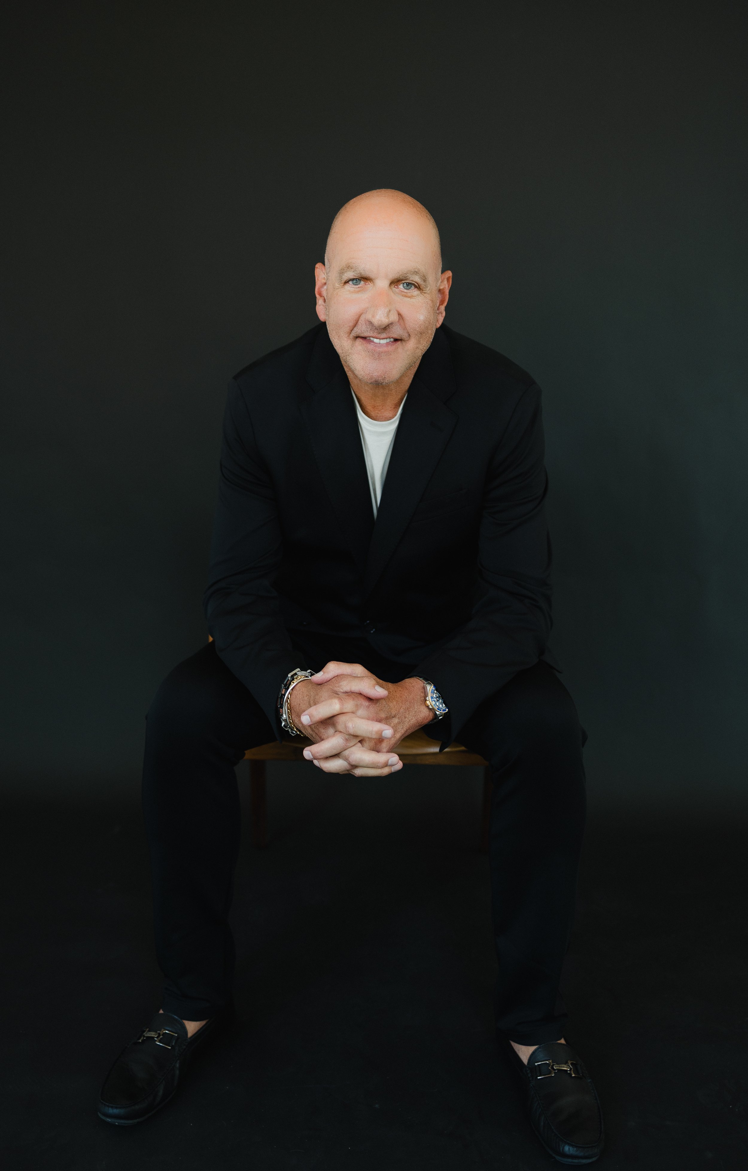 Portrait of a middle-aged bald man with blue eyes, dressed in a black blazer, white shirt, and black shoes, sitting on a wooden chair against a dark gray background, smiling at the camera.