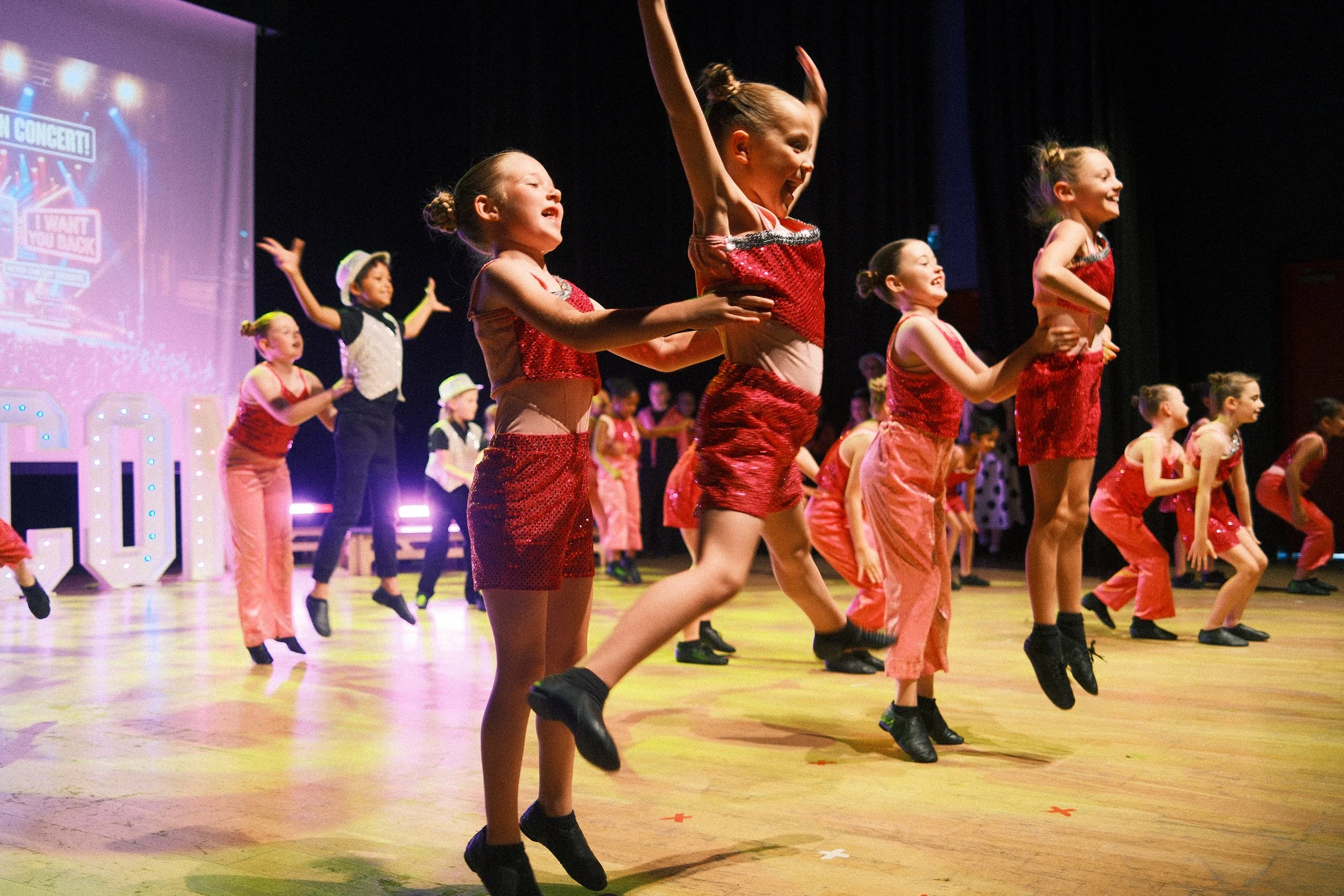 Young girls dressed in red costumes dancing on stage during a performance with musical notes and stage lights in the background.