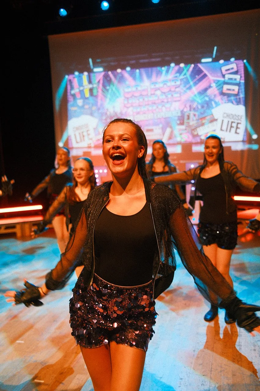 Young woman smiling and dancing on stage with a group of girls during a performance, colorful lights and a vibrant backdrop.