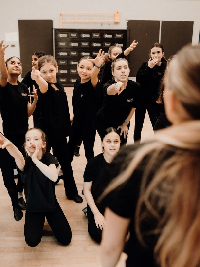 A group of young girls in black clothing participating in a dance or acting class, with some standing and others kneeling, as they listen to or interact with an instructor in a dance studio.