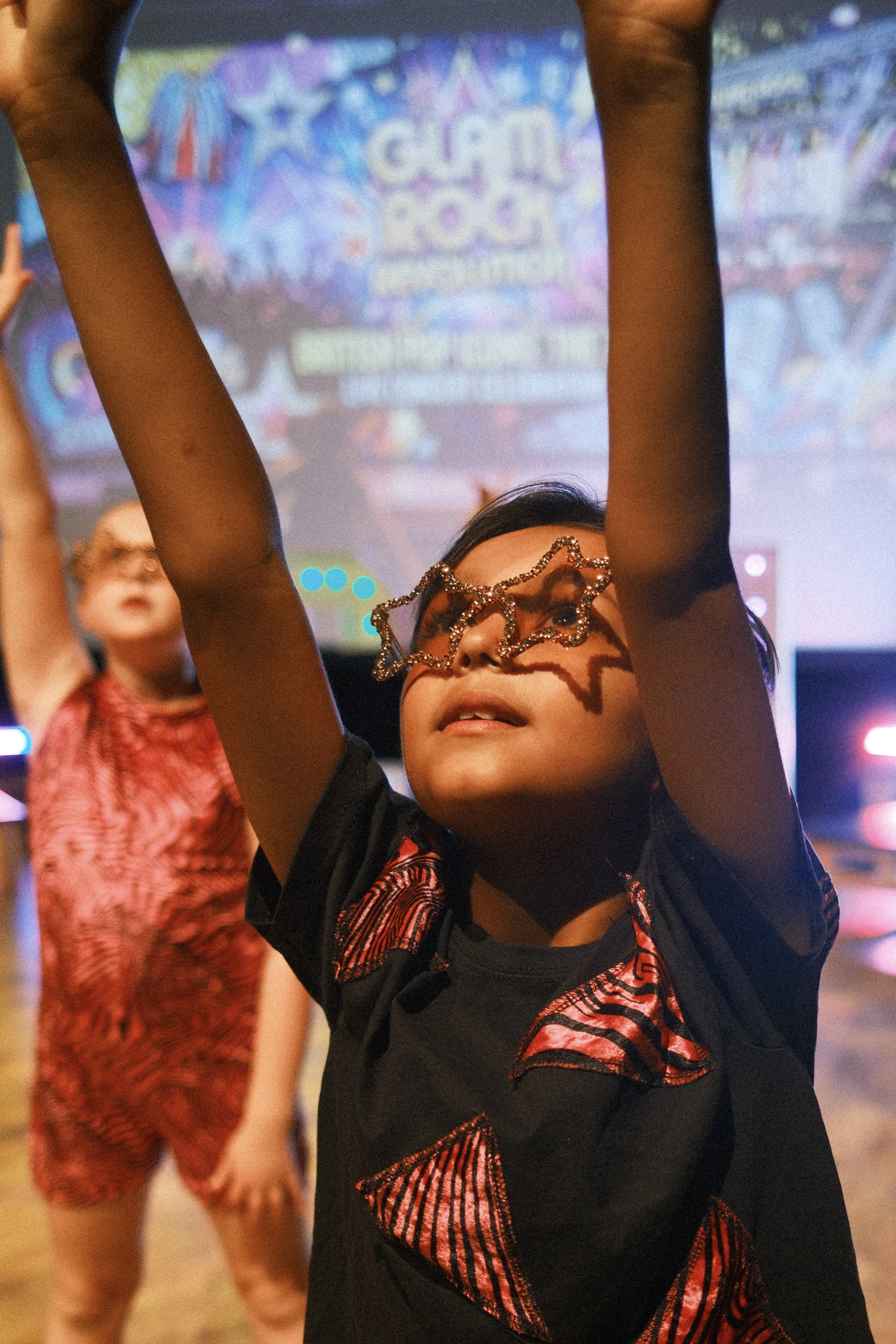 A young girl with dark hair wearing star-shaped glasses decorated with small beads, standing in a colorful indoor setting with other children in the background.