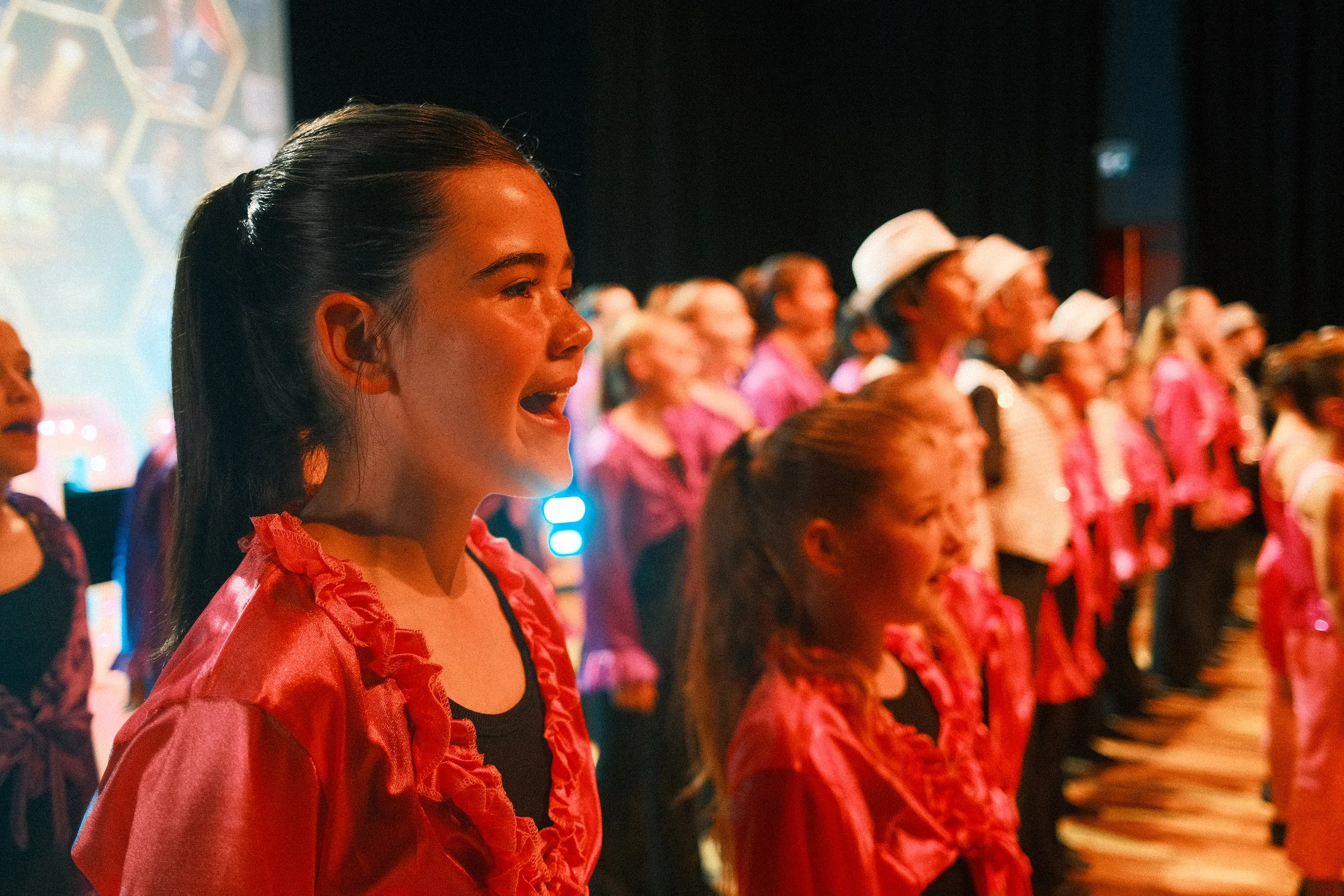A group of children dressed in red and pink shirts standing in a line, singing or performing in a dimly lit indoor setting.