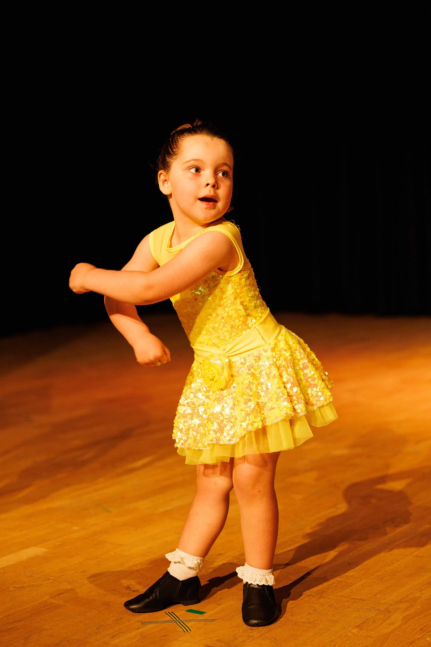 A young girl wearing a yellow dress, black dance shoes, and white socks with lace trim, performing a dance move on a wooden stage with a black background.