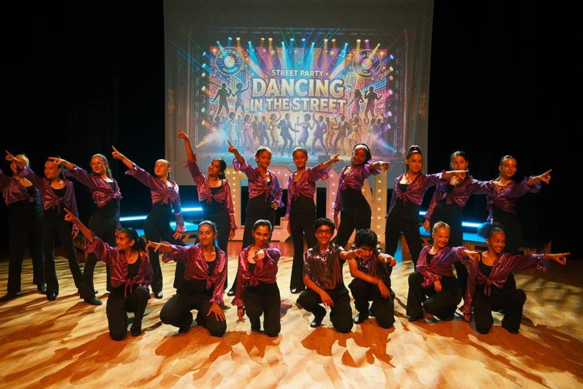A group of young dancers in purple costumes performing on stage during a street party event called 'Dancing in the Street'.