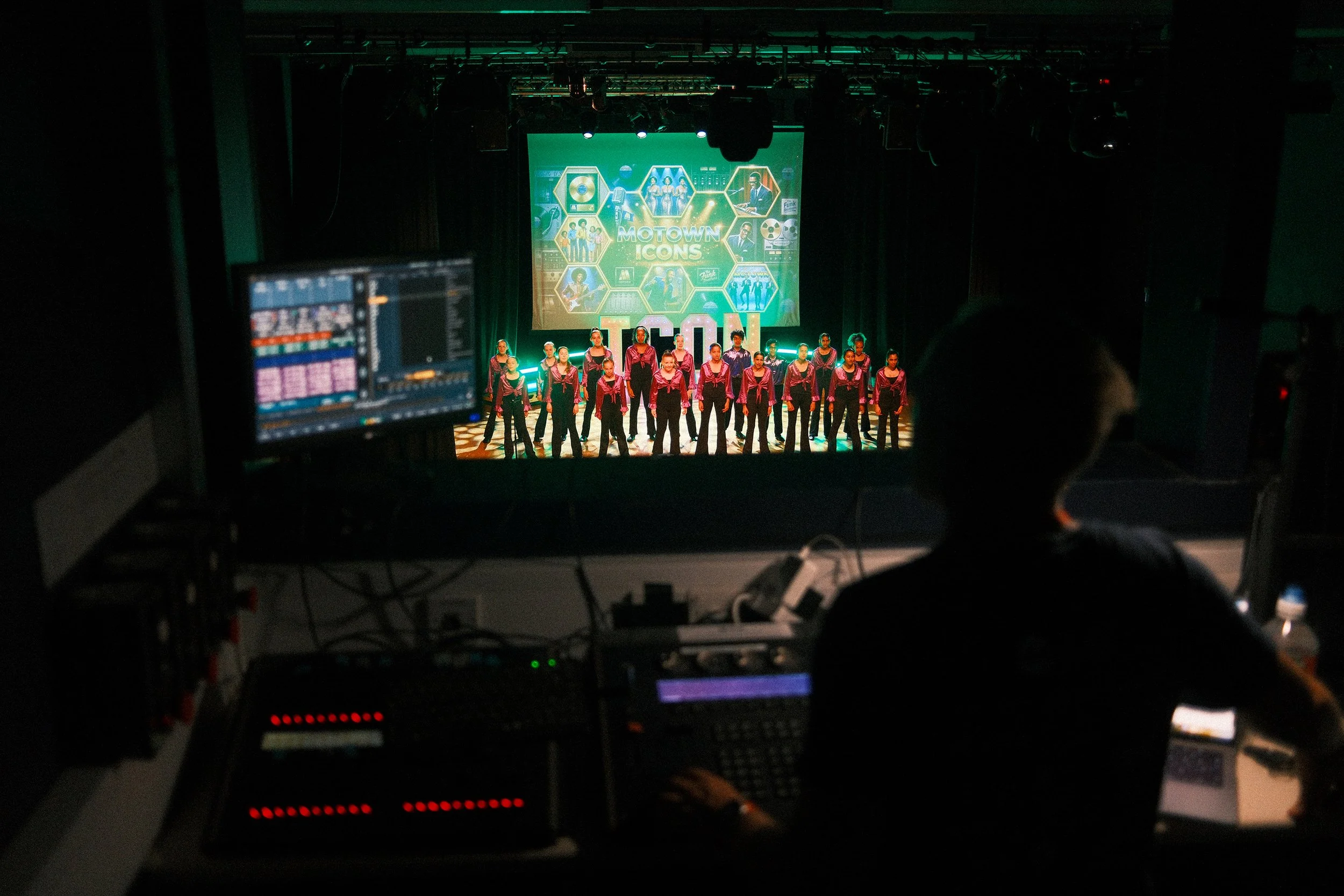 Stage with a group of performers in pink shirts, and a screen displaying the words 'Motown Icons' in the background, viewed from behind the control room with lighting and audio equipment.