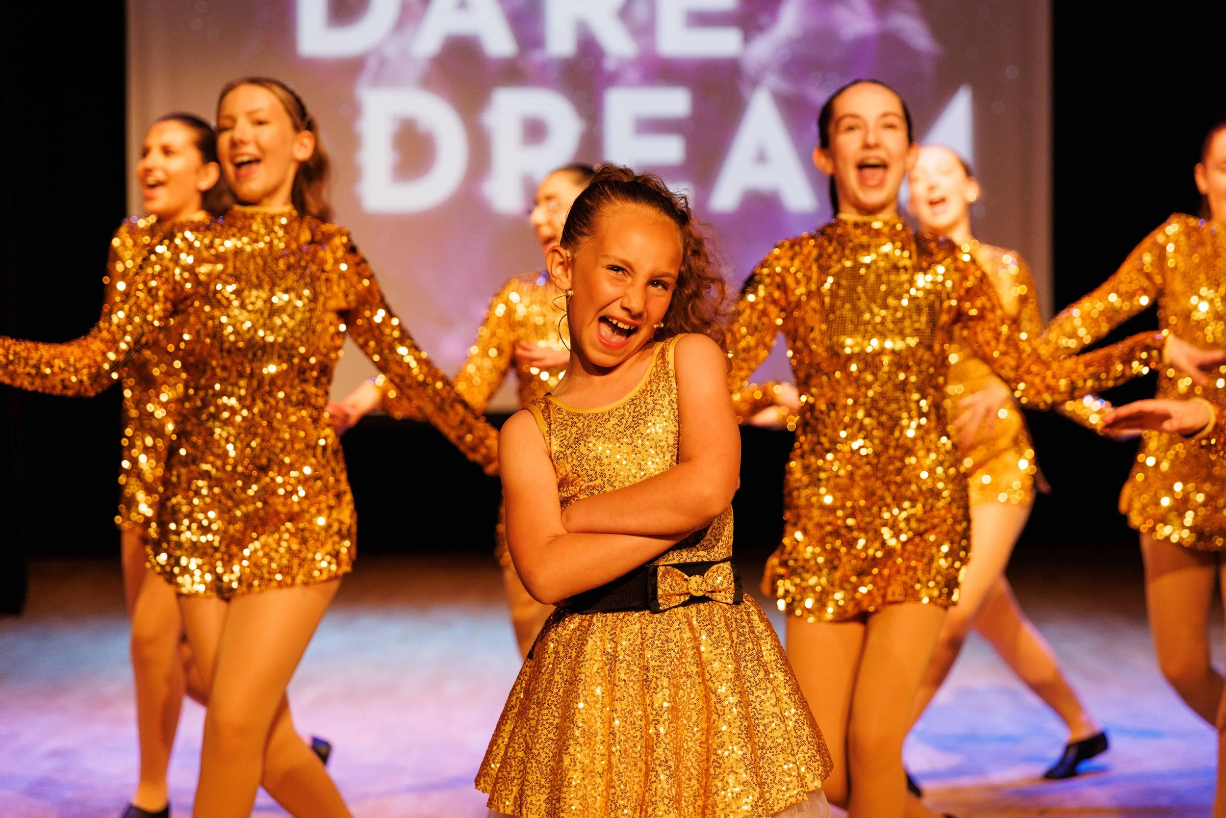 Young girl with curly hair in a gold dress posing confidently on stage during a dance performance, with other dancers in gold costumes behind her.