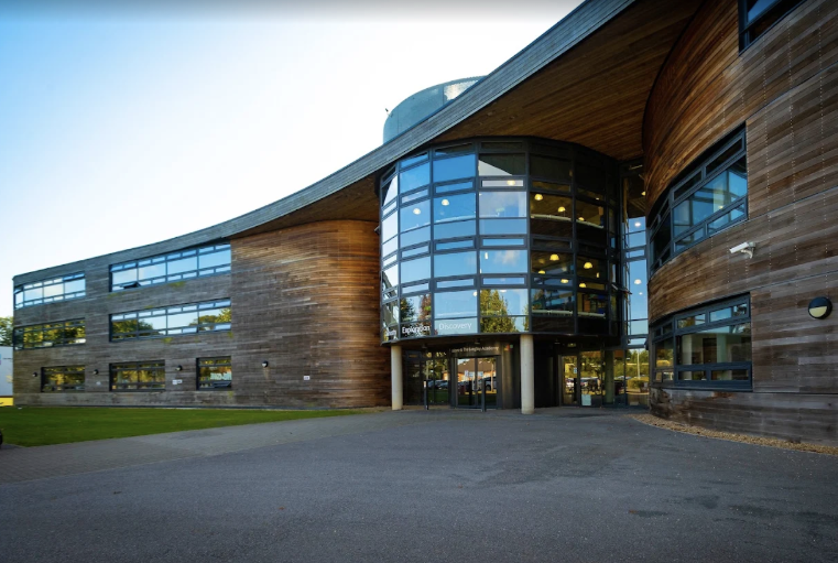 Modern building with curved wooden exterior and large glass windows, surrounded by a paved area and lawn