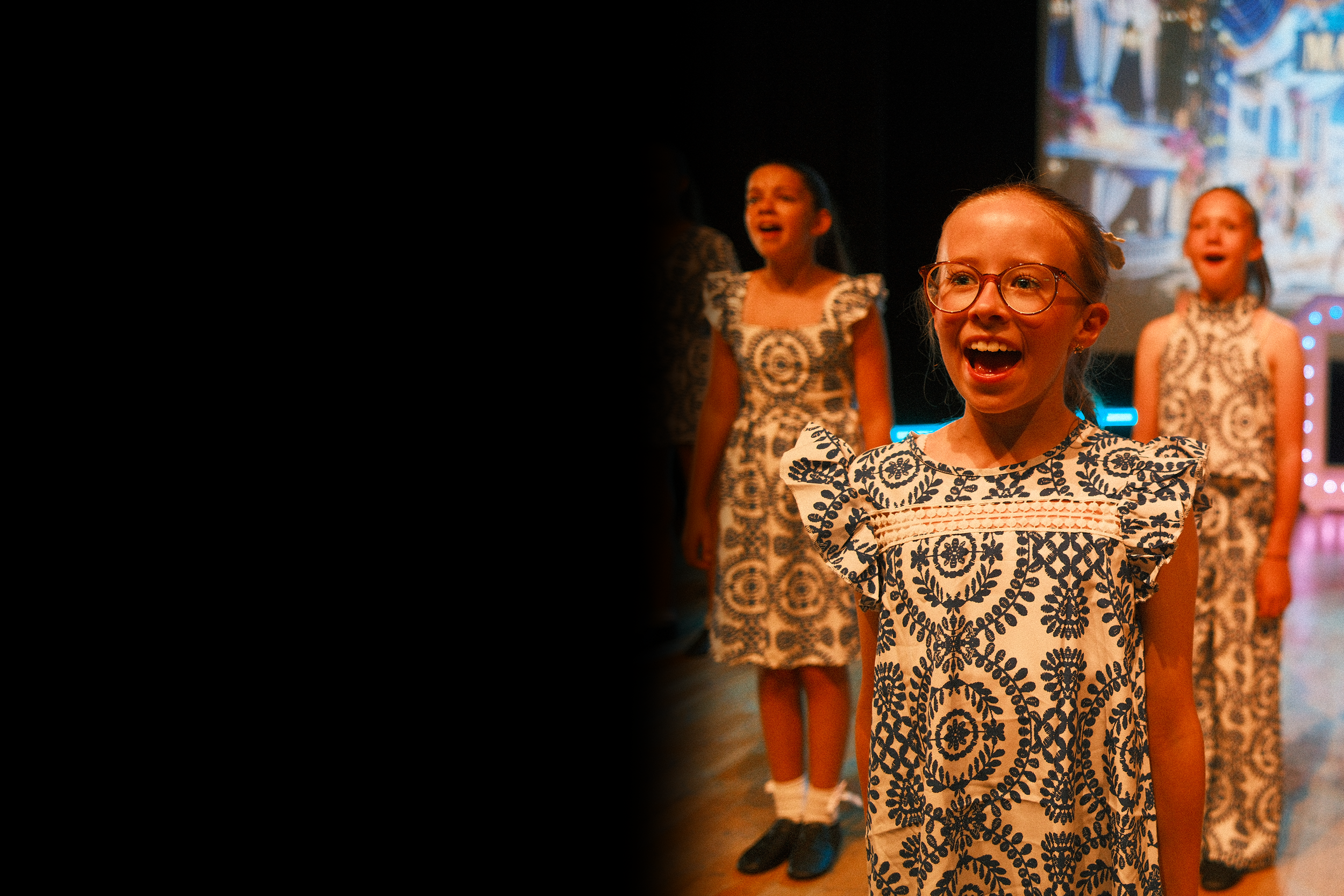 Young girl with glasses singing on stage with other children in the background.