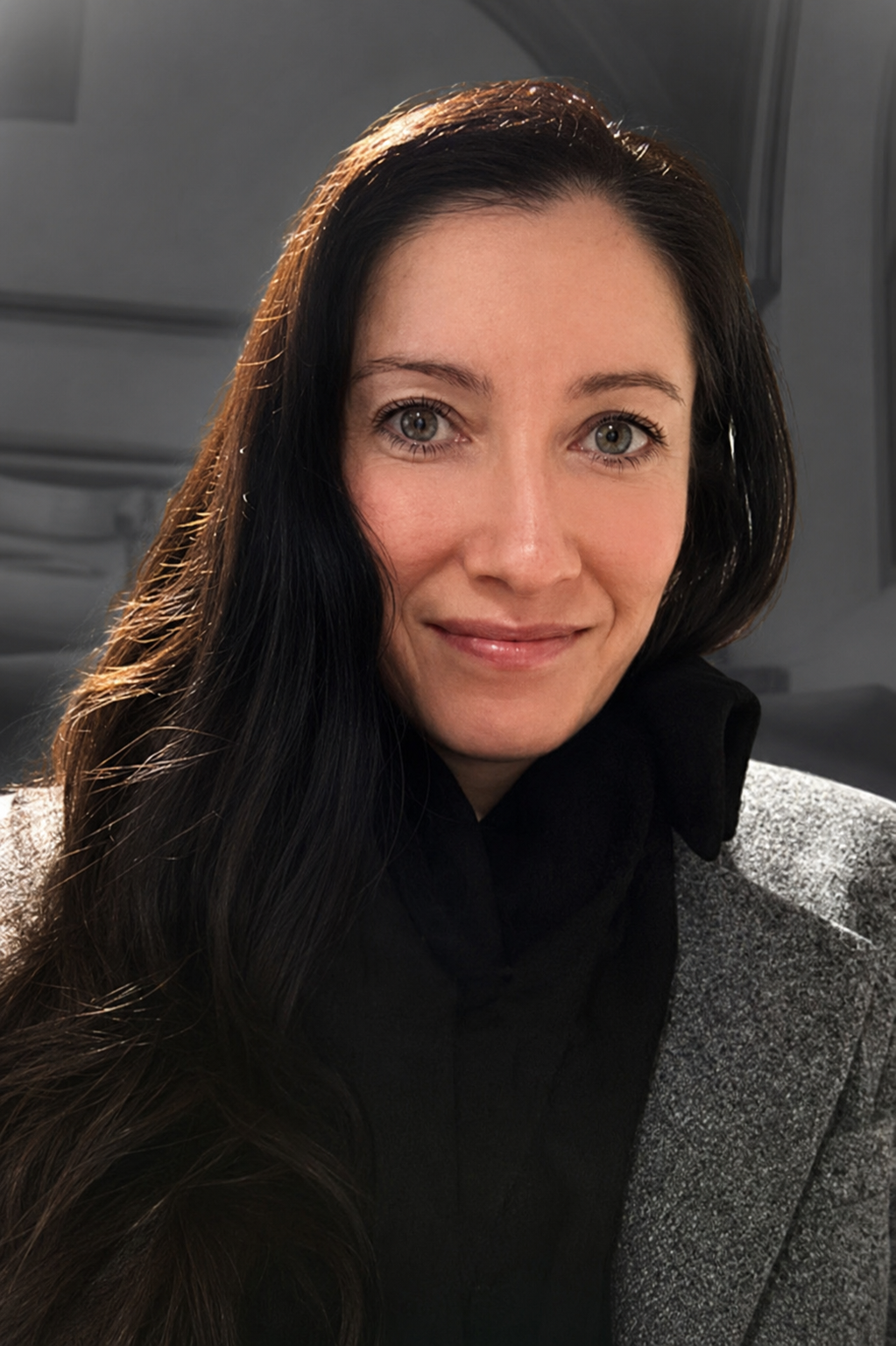 A woman with long dark hair, light skin, and light-colored eyes, smiling at the camera. She is wearing a black top and a gray jacket, and is indoors with a gray background.