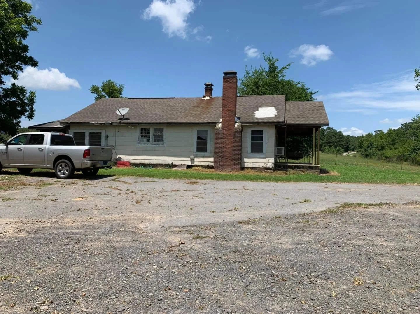 A house with a brick chimney, satellite dish, and air conditioning window units, sitting on a grassy lot with a gravel driveway in front, under a partly cloudy sky.
