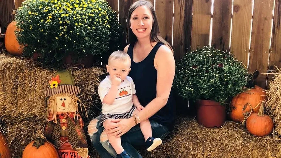 A woman and a young child sitting on hay bales in front of a wooden fence, surrounded by pumpkins and fall decorations, including a scarecrow with a striped hat and a plaid outfit, during an autumn festival.