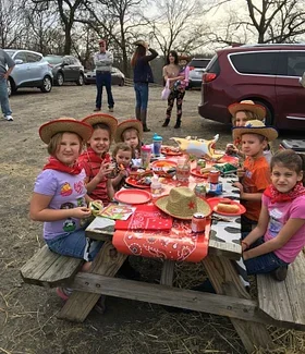 Children having a picnic at a wooden table outdoors with food, drinks, and decorated tablecloth, while adults stand in the background near parked cars.