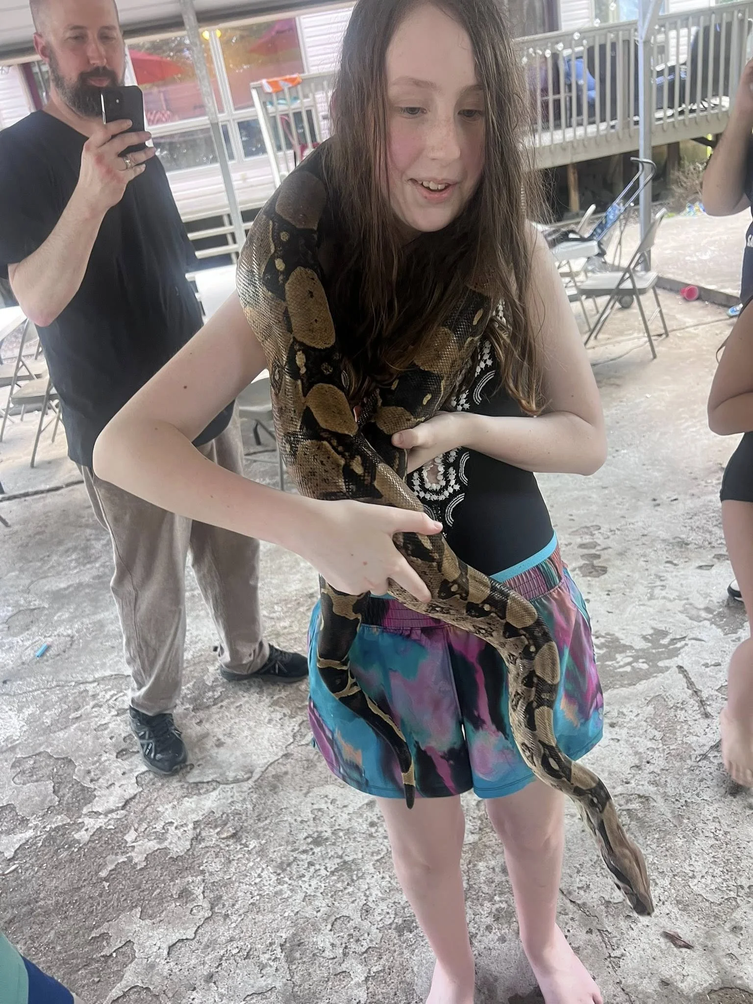 Young woman holding a large snake on her arm, on a cracked concrete surface in an outdoor setting with chairs and a railing in the background.