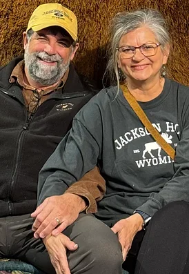 A smiling man and woman sitting close together, holding hands, with a wooden background. The man is wearing a yellow hat and a black jacket, and the woman is wearing glasses and a gray sweatshirt with 'Jackson Hole Wyoming' written on it.