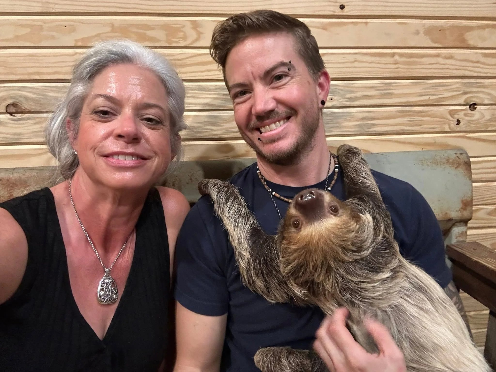 A smiling older woman with gray hair and a necklace taking a selfie with a younger man with short brown hair, earrings, and beaded necklaces. They are indoors sitting against a wooden wall with a dog in the younger man's arms.
