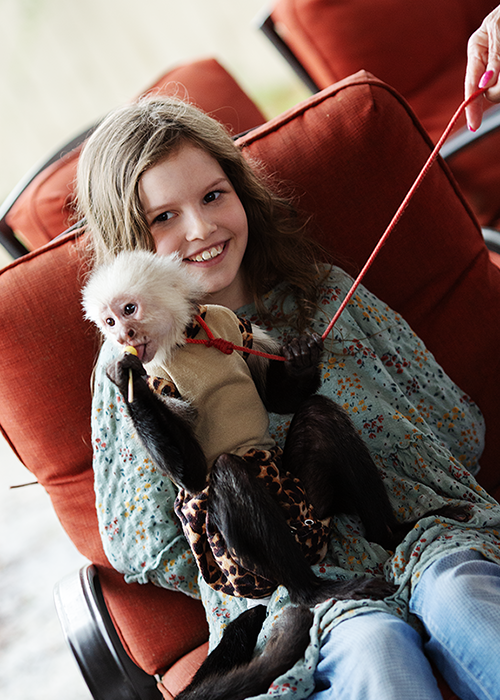 A young girl sitting on a red armchair, holding a small dog dressed in a dog costume, smiling at the camera.