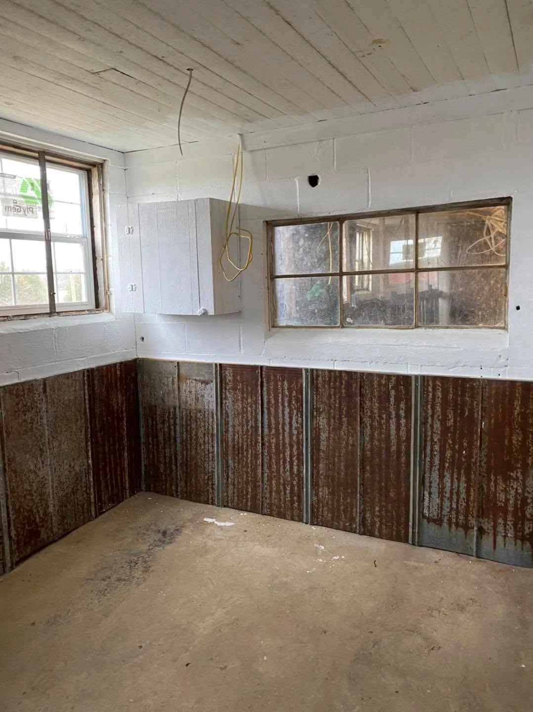 Interior of a room under construction with exposed wiring, unpainted drywall, and metal wall panels, with high windows and a concrete floor.