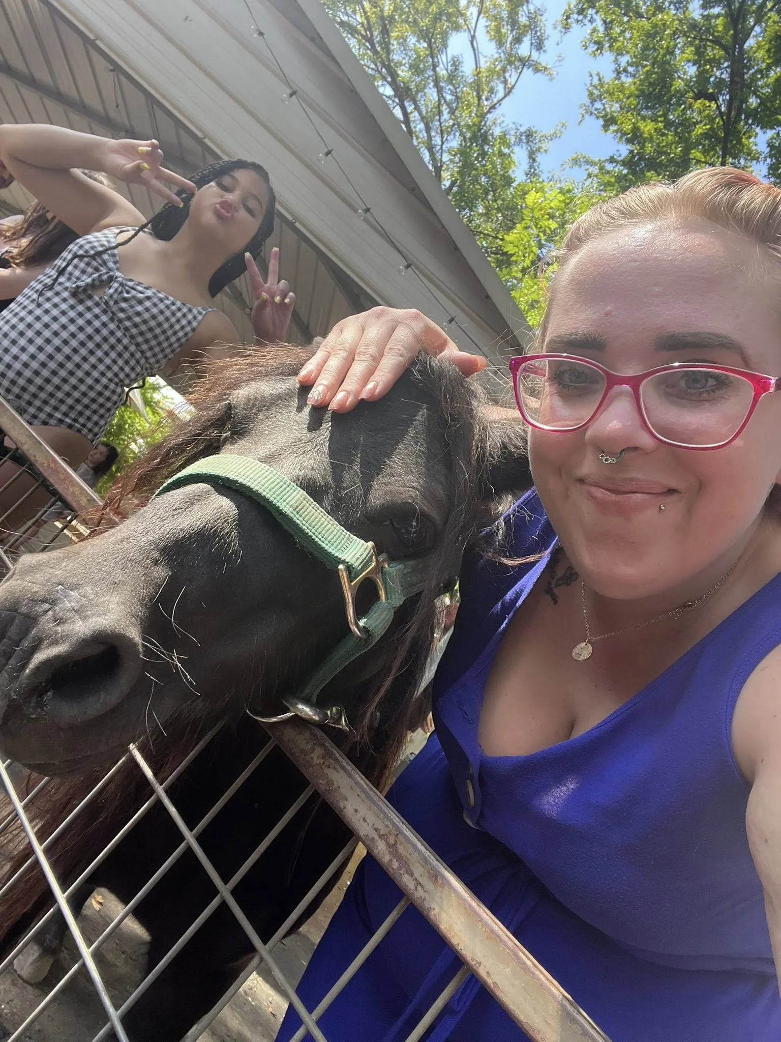 Two women at a farm or petting zoo, one petting a small horse or pony, the other making a peace sign, outdoors with trees and a building overhead.