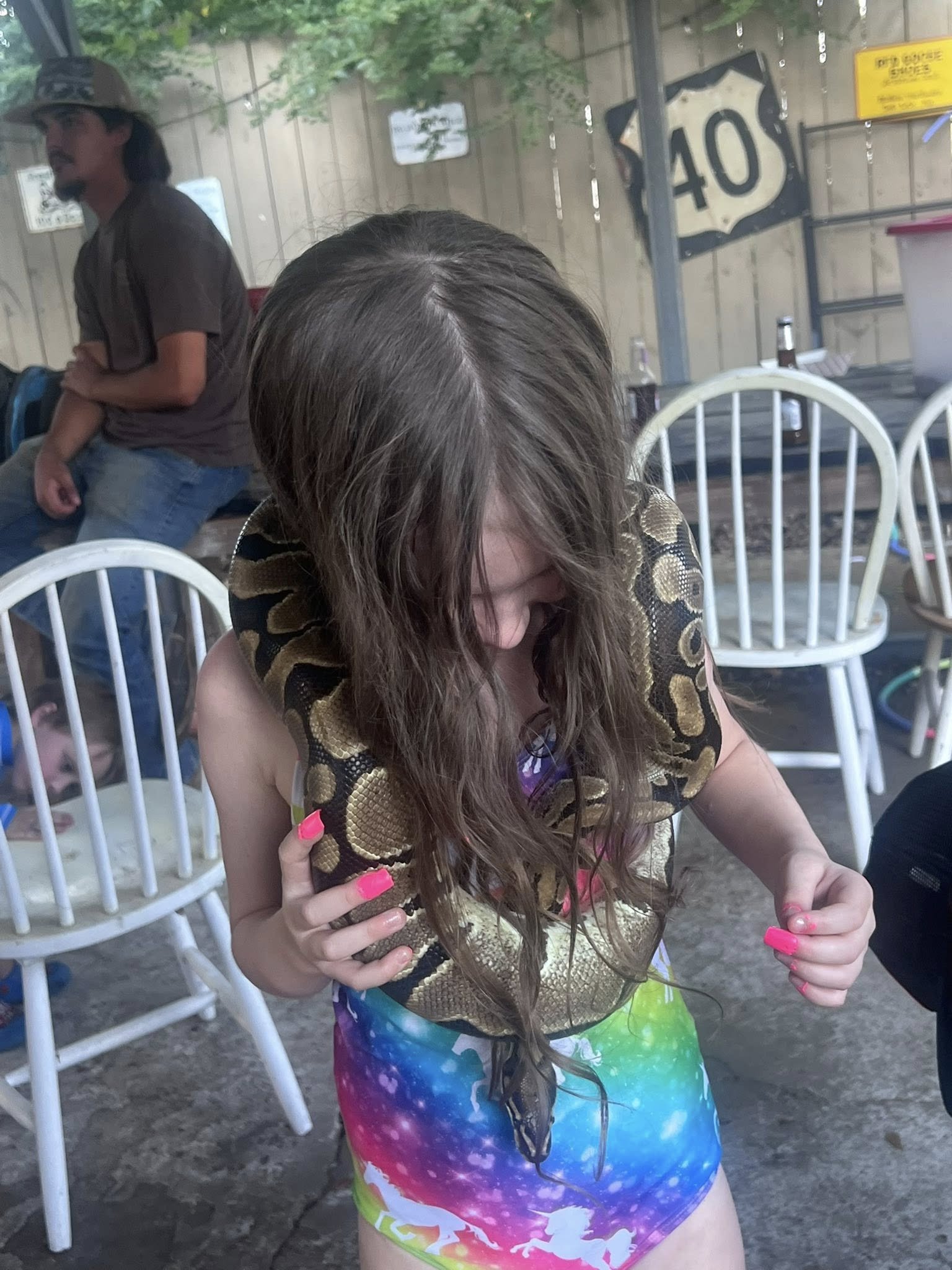 A young girl with long brown hair wearing a colorful tie-dye dress is holding a large snake, which is wrapped around her arm and hand, indoors or on a porch. There are white chairs and bottles in the background, along with a wooden fence and a sign t