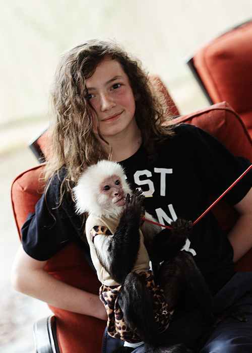 Teen girl with curly hair sitting on a red chair holding a small white monkey wearing a tan and leopard print outfit.