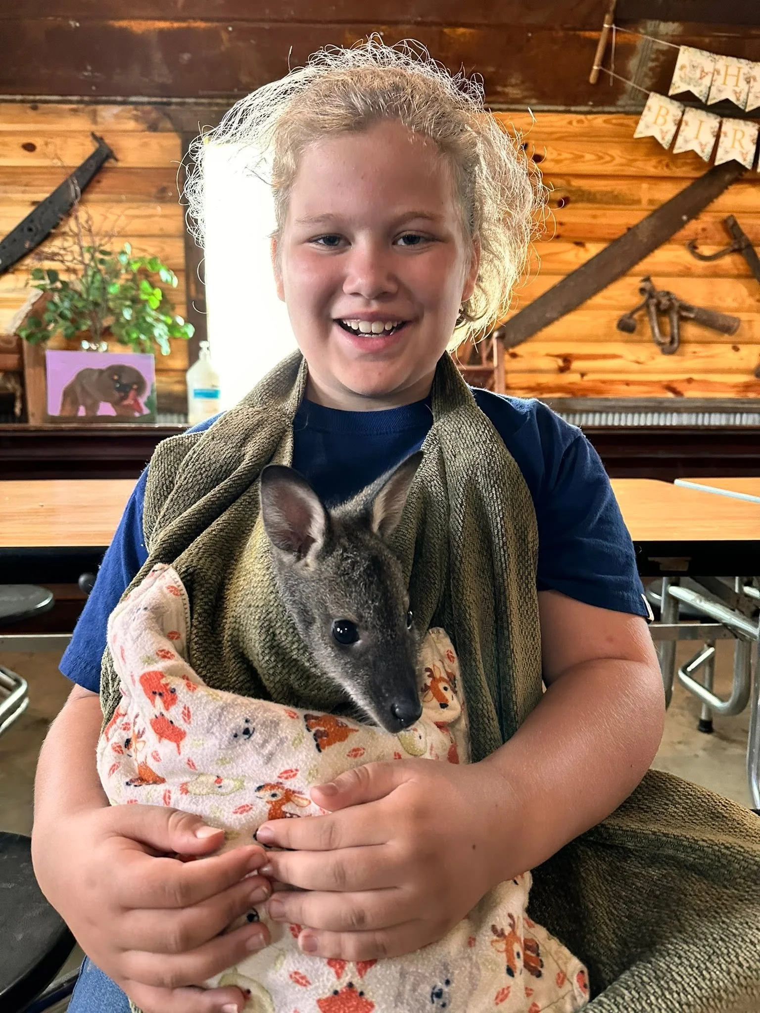 A smiling young girl with curly blonde hair holding a small, grey and white puppy wrapped in a festive blanket. They are inside a rustic wooden room with vintage tools and a 'Happy Birthday' banner in the background.