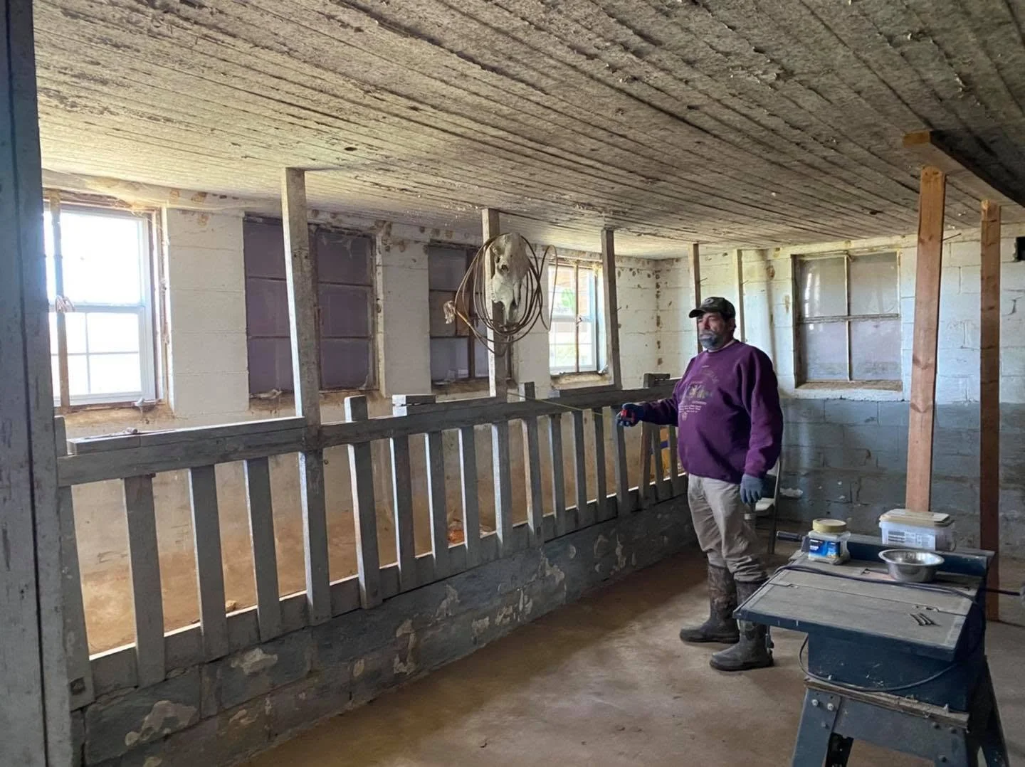 A man in a purple sweatshirt and boots standing inside a house under construction, holdign a tool in his right hand, with a star-shaped metal object hanging from a wire above him and several windows in the background.