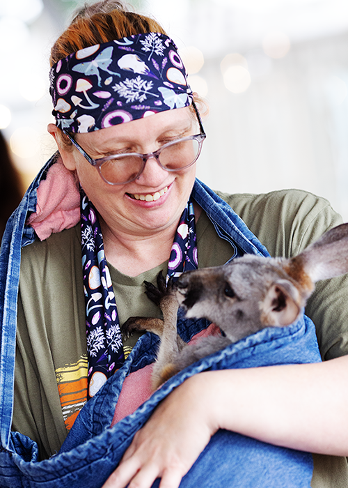 A woman with red hair, wearing glasses, a colorful bandana, and a green shirt, smiling while holding a small kangaroo in a blue pouch