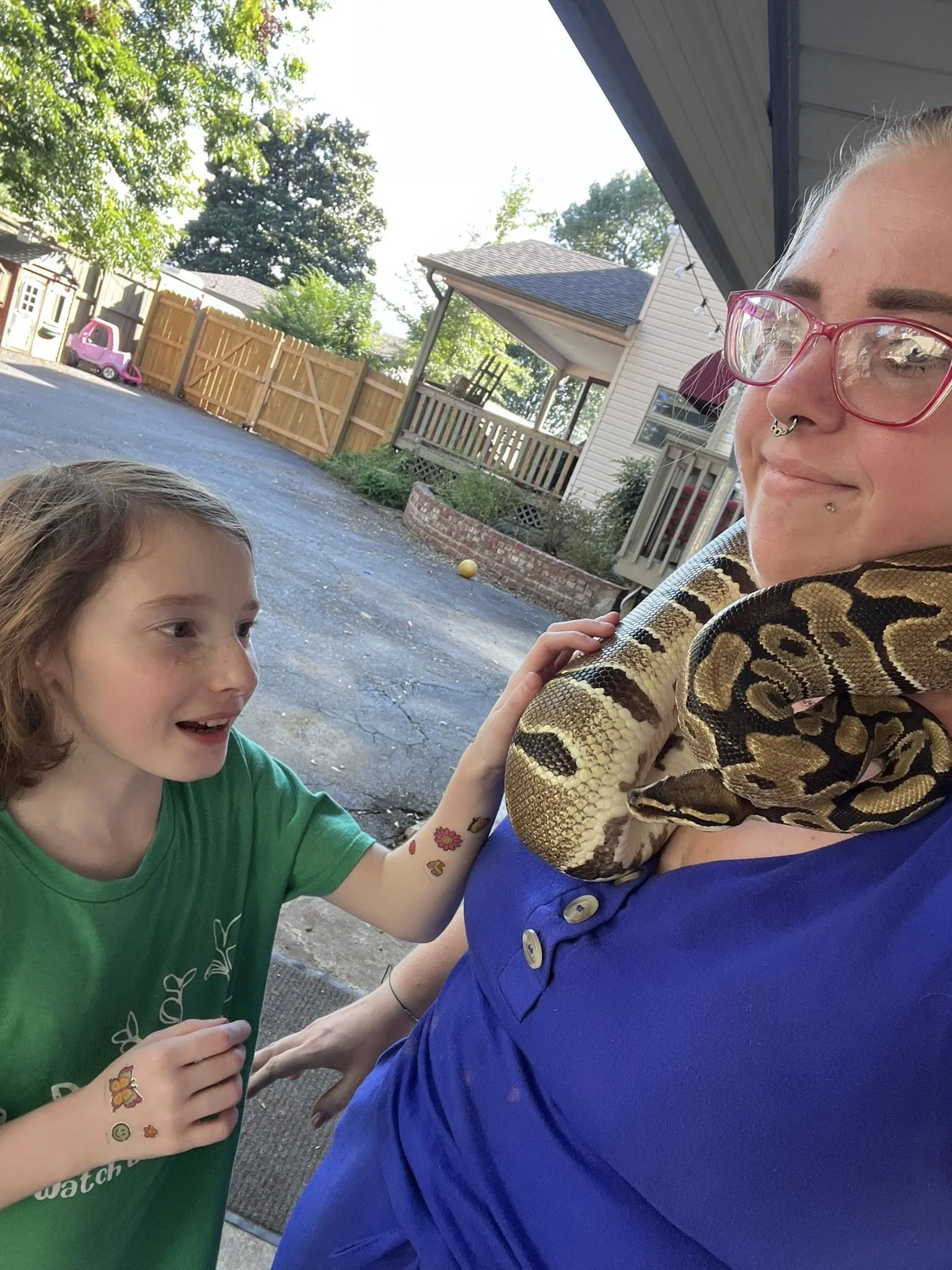 A young girl with tattoos on her arm petting a large snake being held by a woman in a blue shirt and pink glasses outside in a backyard.