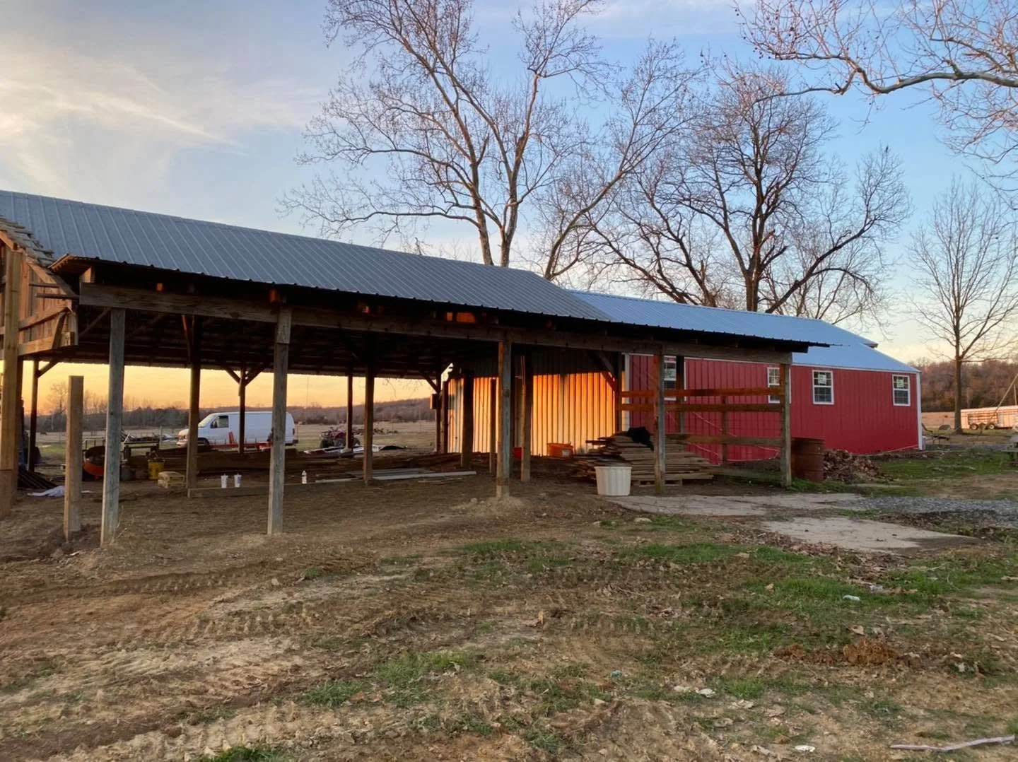 A red barn with a metal roof under construction, surrounded by trees and open land during sunset.